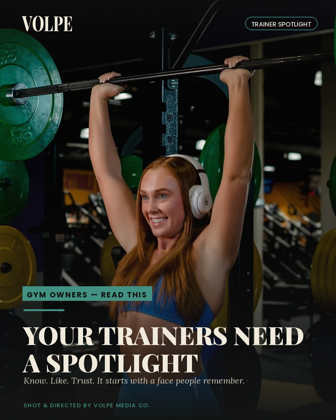 A woman with long red hair wearing headphones, smiling, lifting a barbell overhead in a gym, with weight plates visible in the background.