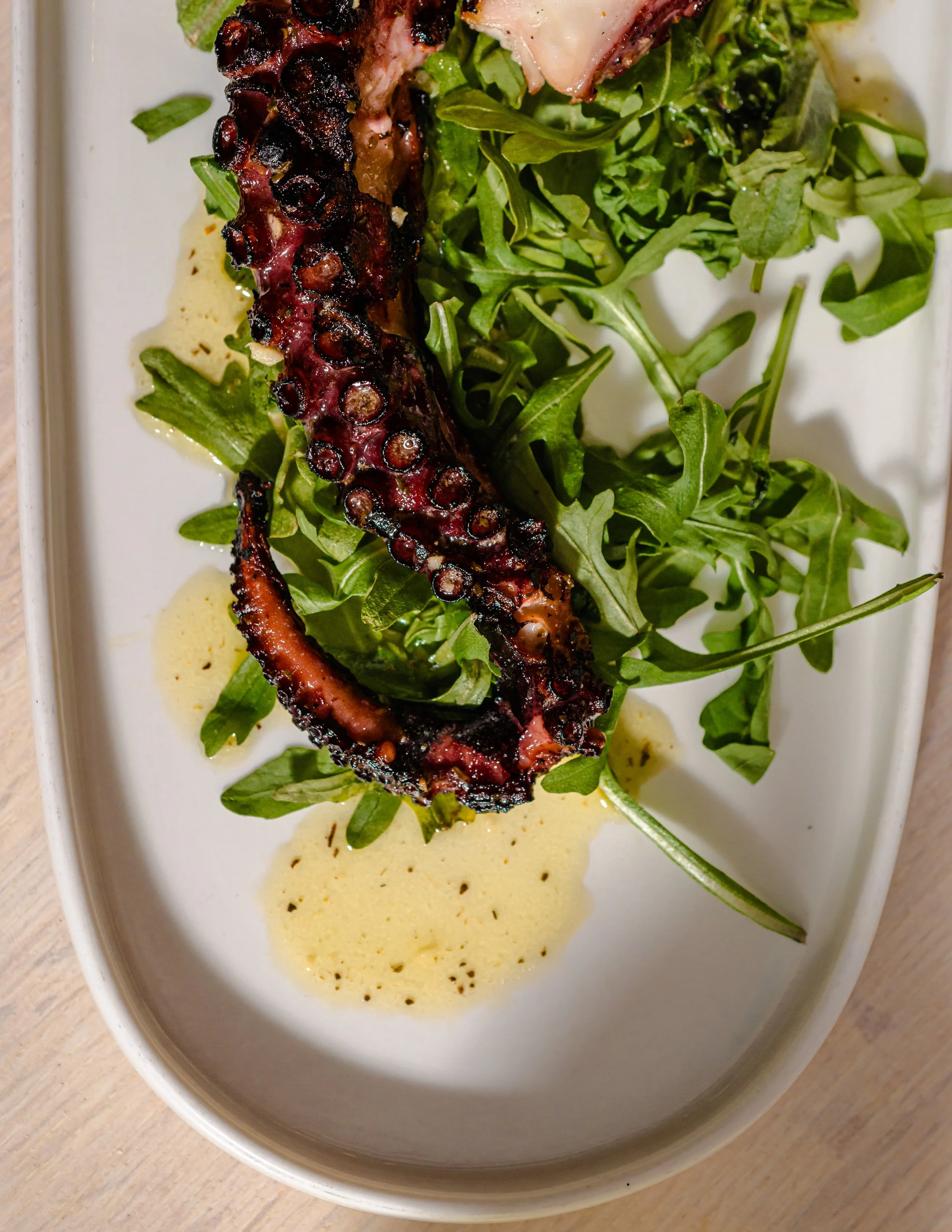 A grilled octopus tentacle served with fresh arugula on a white rectangular plate with a creamy yellow sauce.