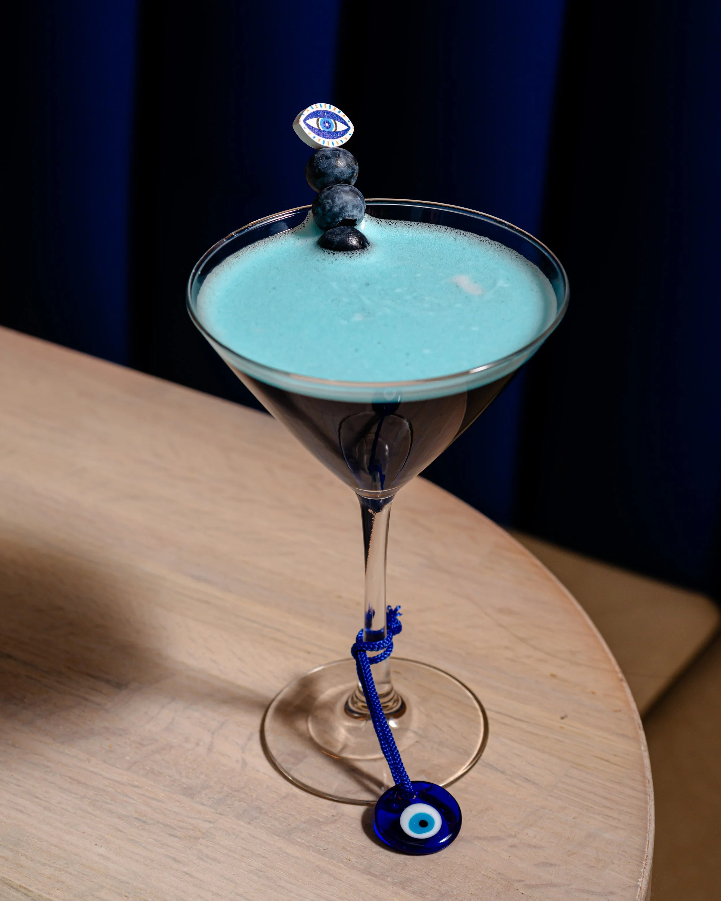 A fancy cocktail glass filled with a blue frothy drink, topped with four blueberries, a small decorative eye on top, and a blue eye charm hanging from the rim.
