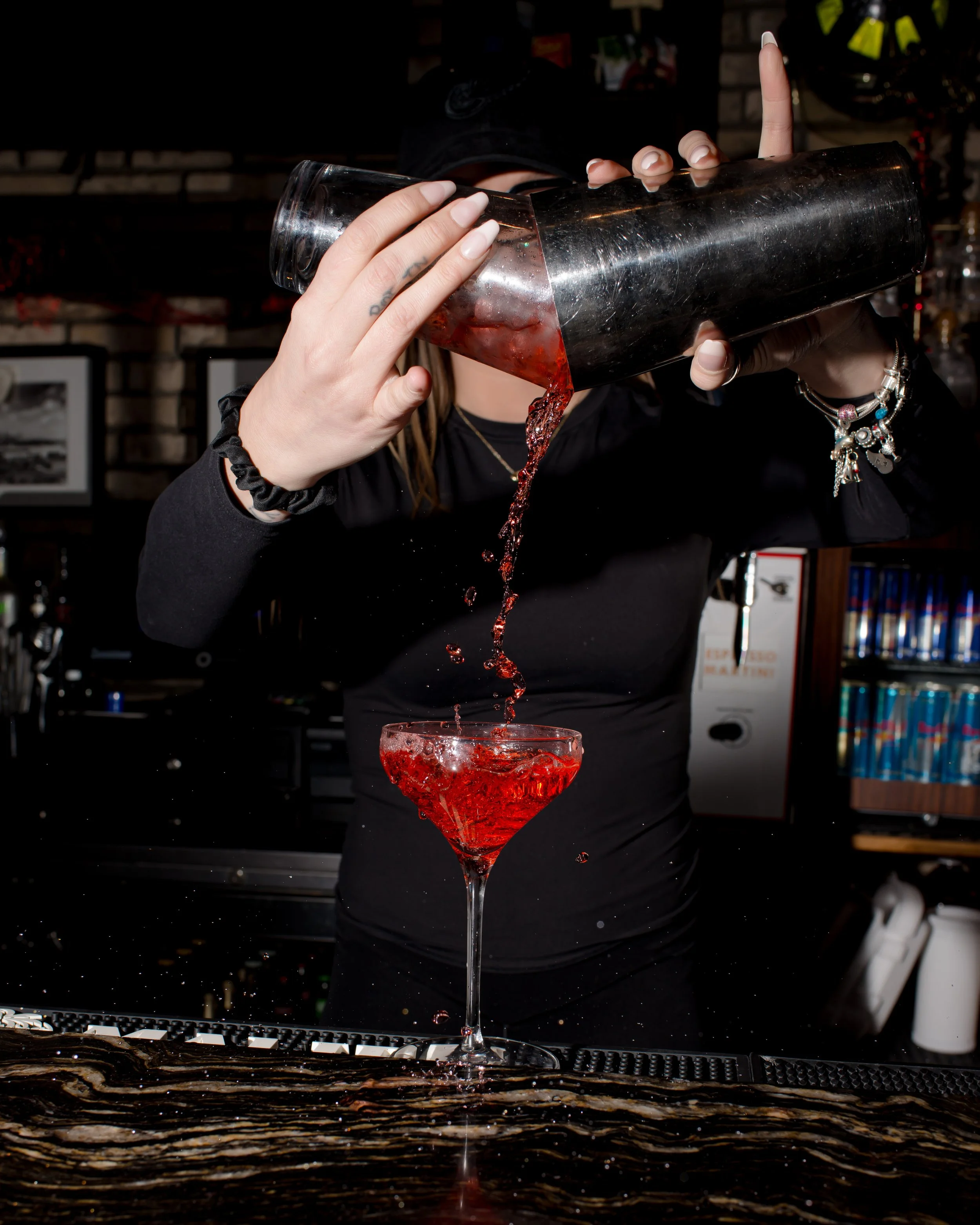 A person pouring red liquid from a cocktail shaker into a martini glass, with liquid splashing and droplets in the air.