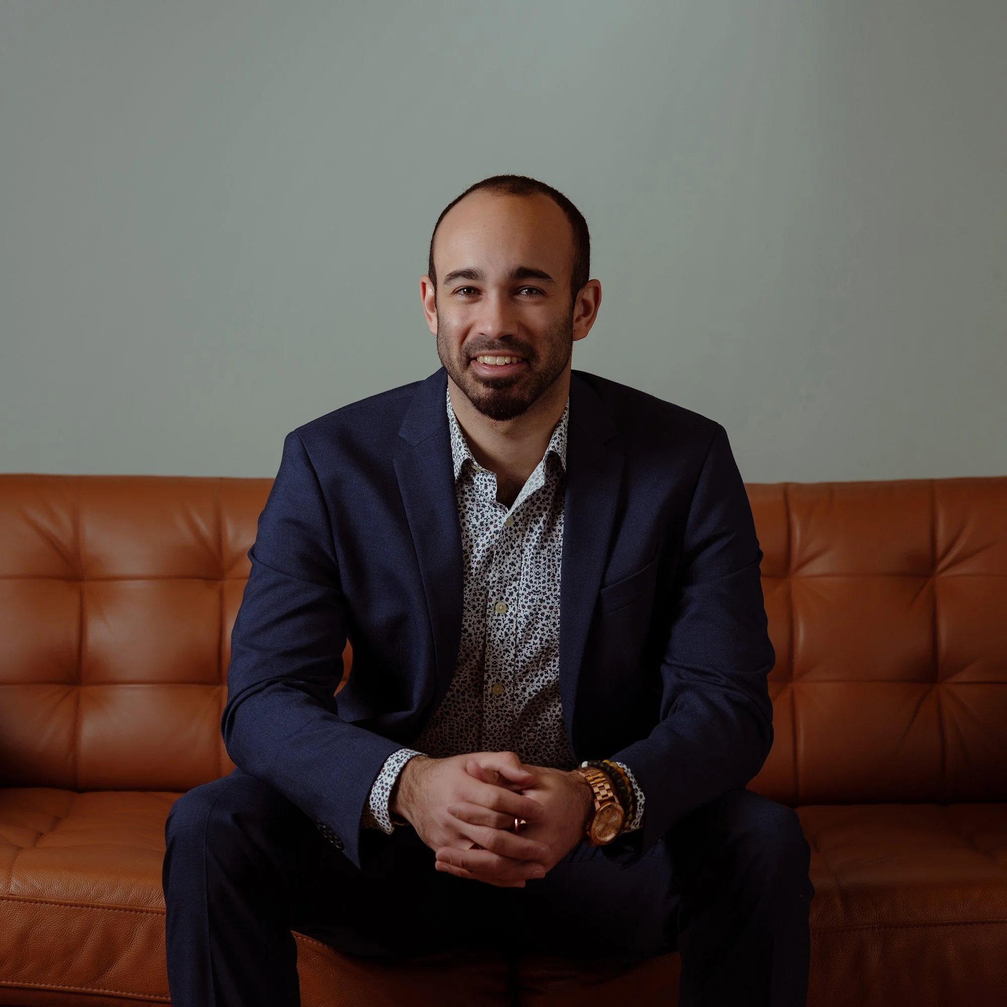 A man with a beard and short dark hair, wearing a navy blazer and a patterned shirt, sitting on a tan leather couch in front of a plain light green wall.