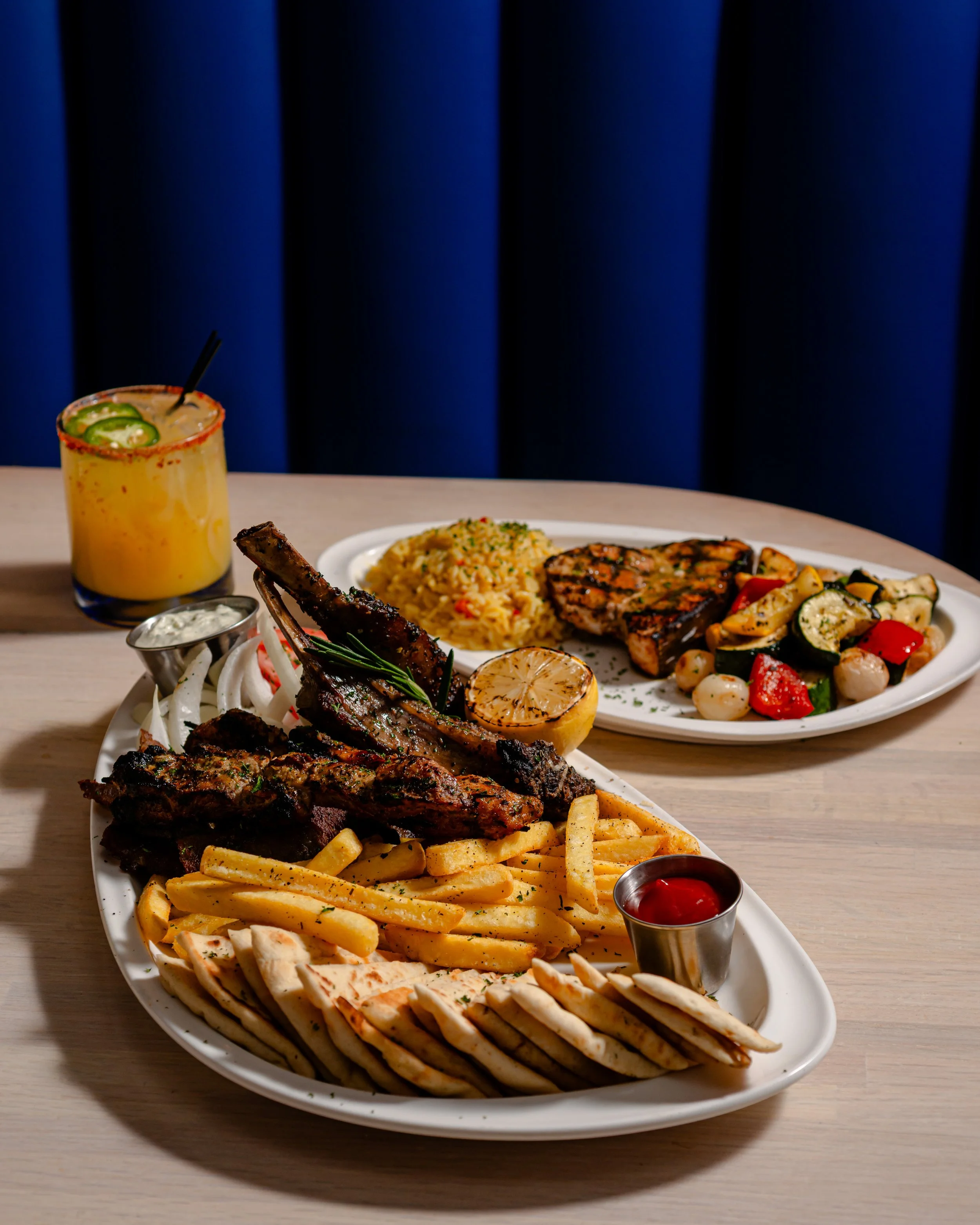 A meal at a restaurant featuring two white oval plates with chicken shawarma, French fries, and pita bread; another plate with rice, grilled chicken, and vegetable salad; a glass of yellow beverage with lime slices and black straw; small containers o