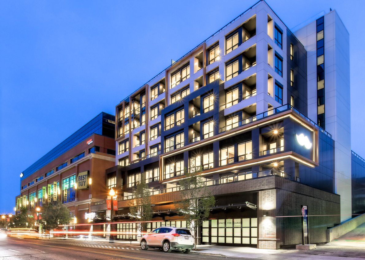 A modern multi-story building with illuminated windows and a prominent Apple logo on the corner, located on a busy city street at dusk, with cars passing by and mix of retail and residential spaces.