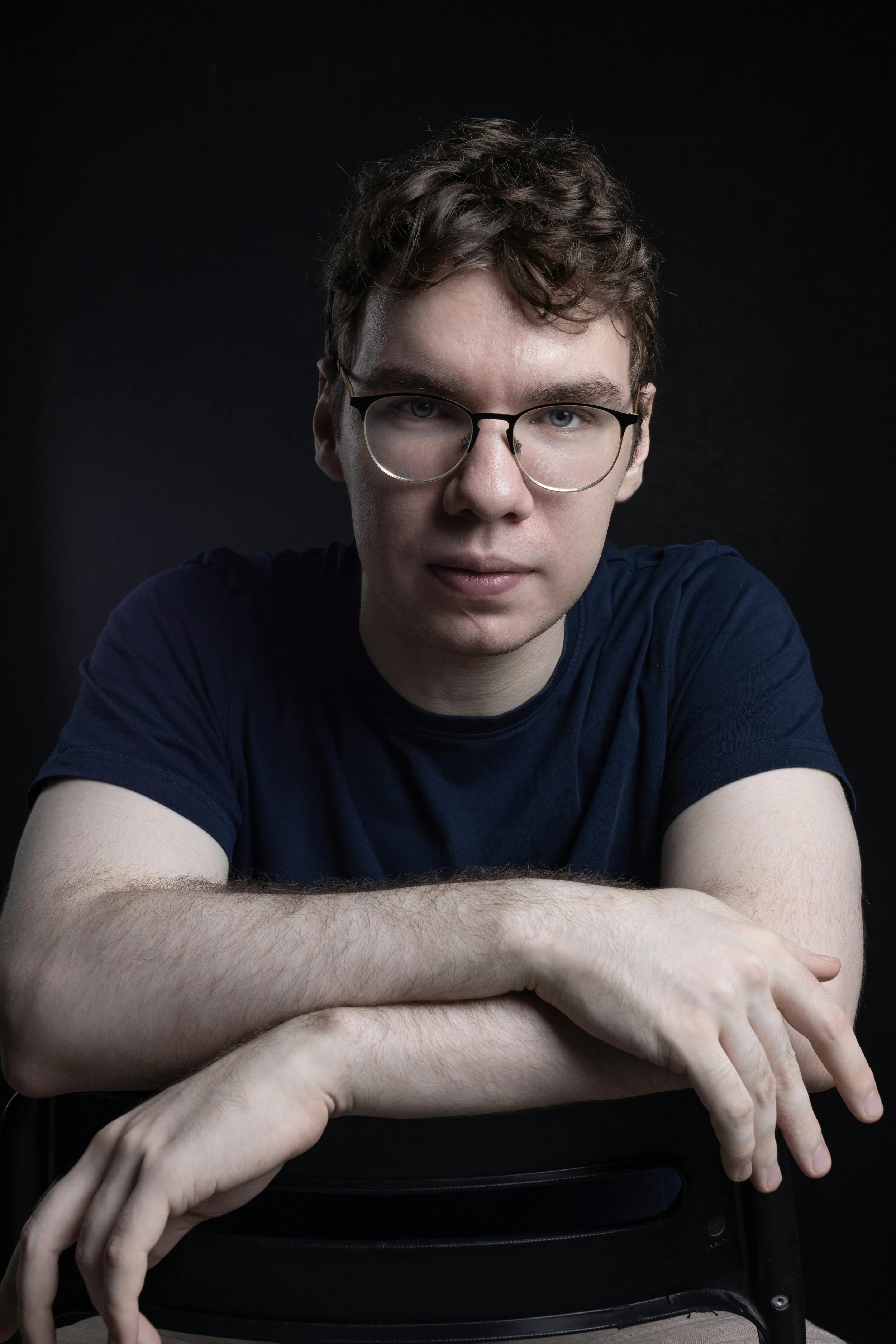 A young man with curly brown hair wearing glasses and a dark blue t-shirt is sitting with his arms crossed on a chair against a black background.