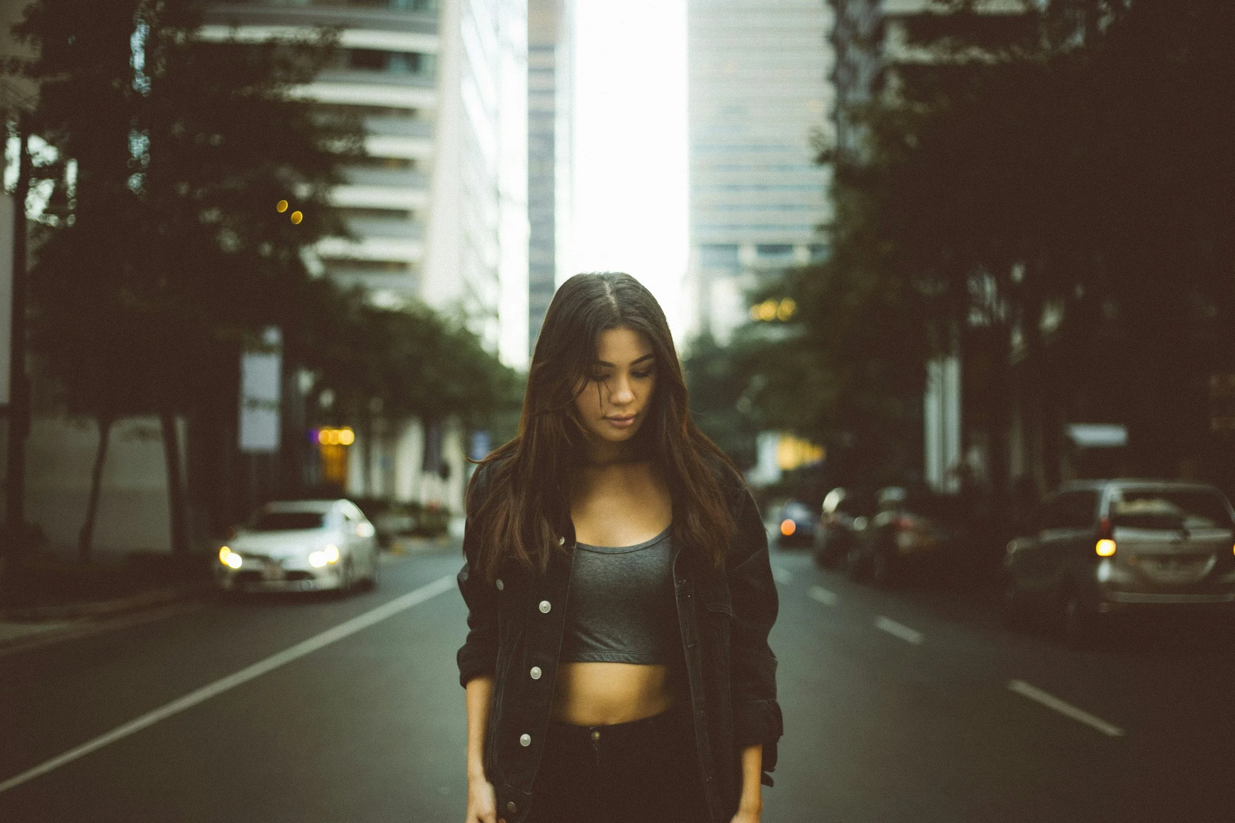 A young woman with long brown hair walking down a city street during the daytime, flanked by tall buildings and parked cars. She is looking down and wearing a black jacket and a gray crop top.
