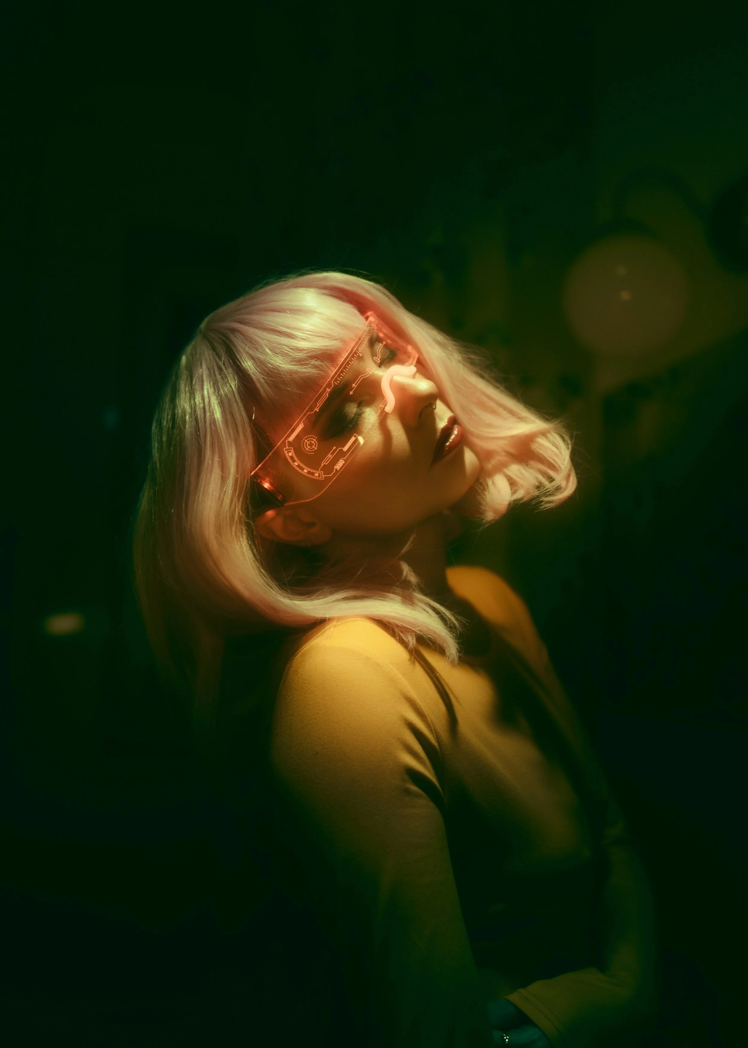 A woman with platinum blonde hair and dark eye makeup is wearing futuristic glasses with holographic displays. She is in a dimly lit environment with a dark green background.