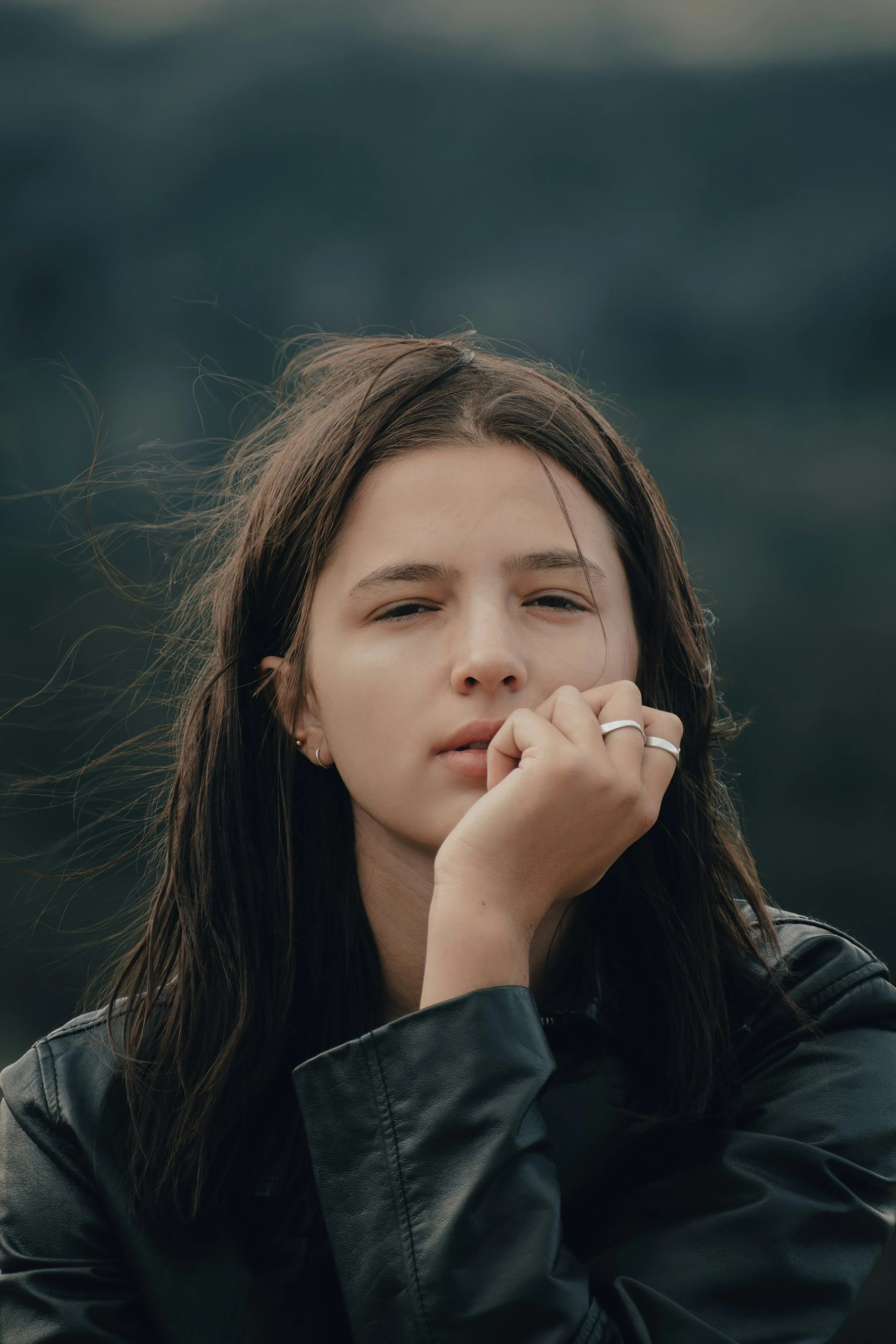 Close-up of a young woman with long brown hair, wearing a black leather jacket, with a contemplative expression, outdoors with a cloudy sky in the background.
