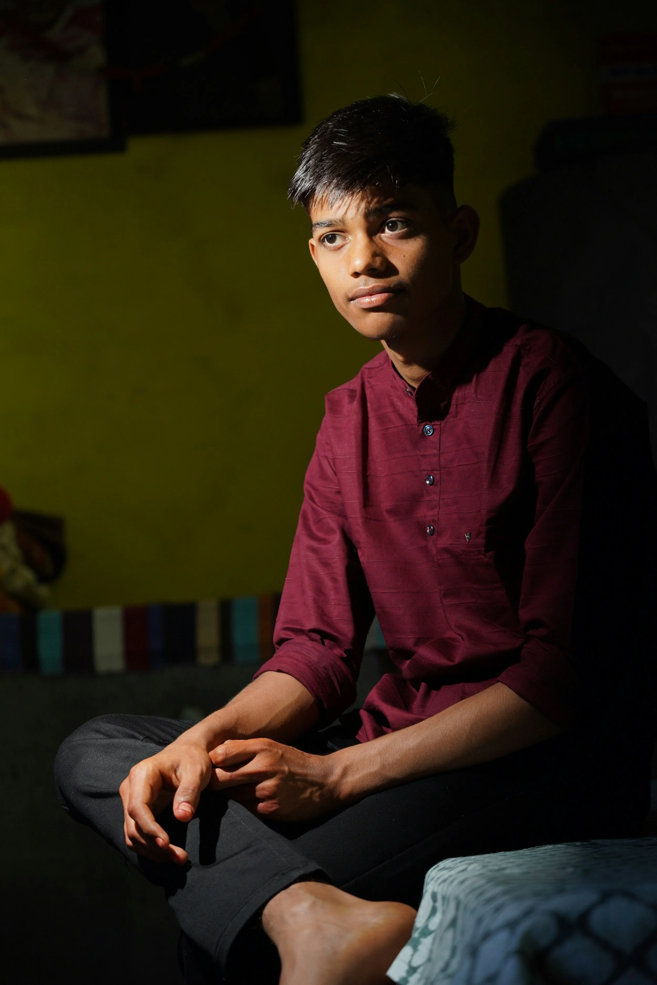 A young man with short dark hair, wearing a maroon shirt, sitting indoors in a shadowed room with a green wall and a black speaker behind him, gazing thoughtfully.