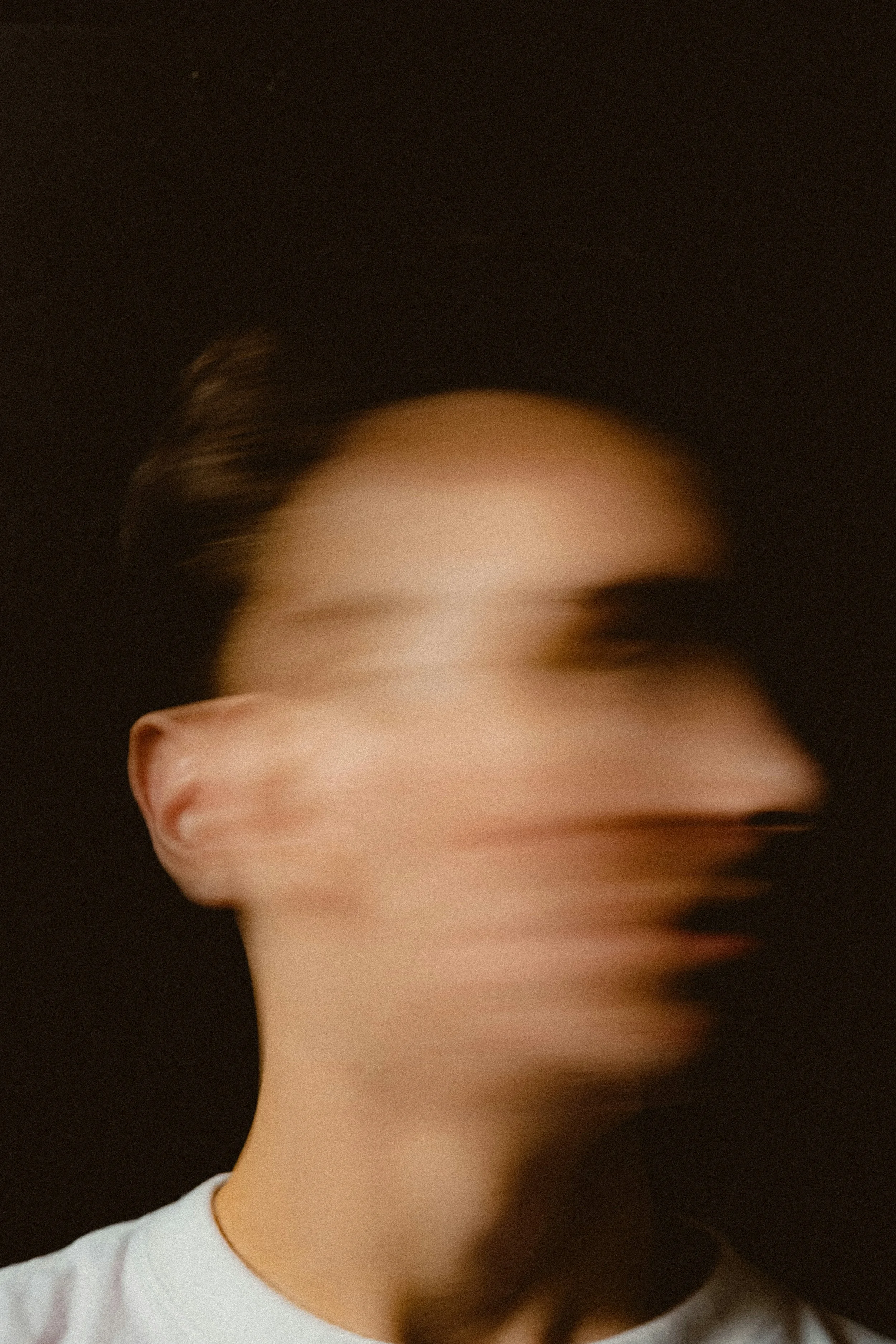 A blurred image of a person with short, brown hair and light skin, wearing a white shirt, against a dark background.