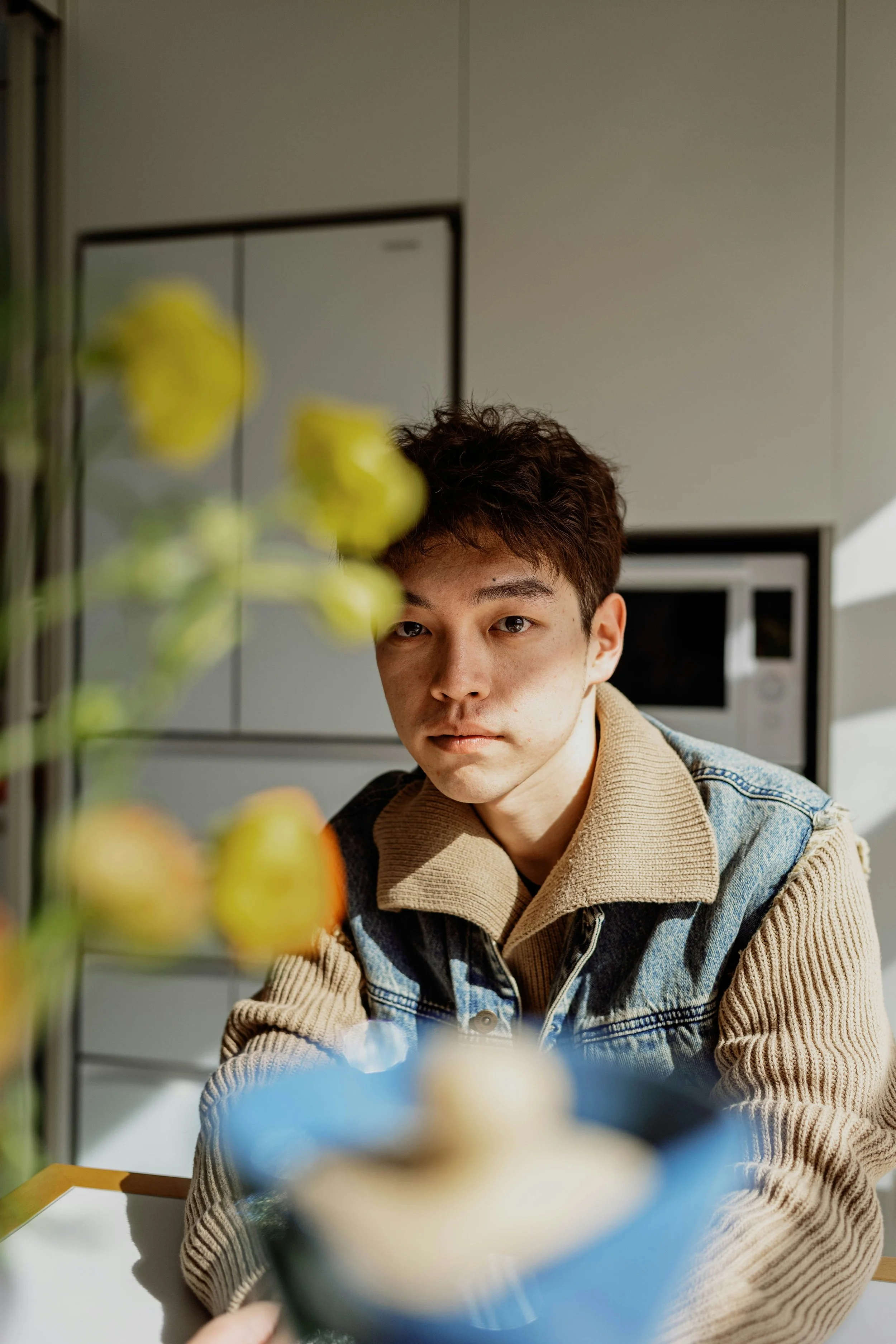 A young man with brown hair and tan skin is sitting at a table, looking at the camera. He is wearing a beige and denim jacket. The image is taken indoors, with a gray wall and kitchen appliances in the background. Bright yellow flowers are in the foreground, slightly out of focus.