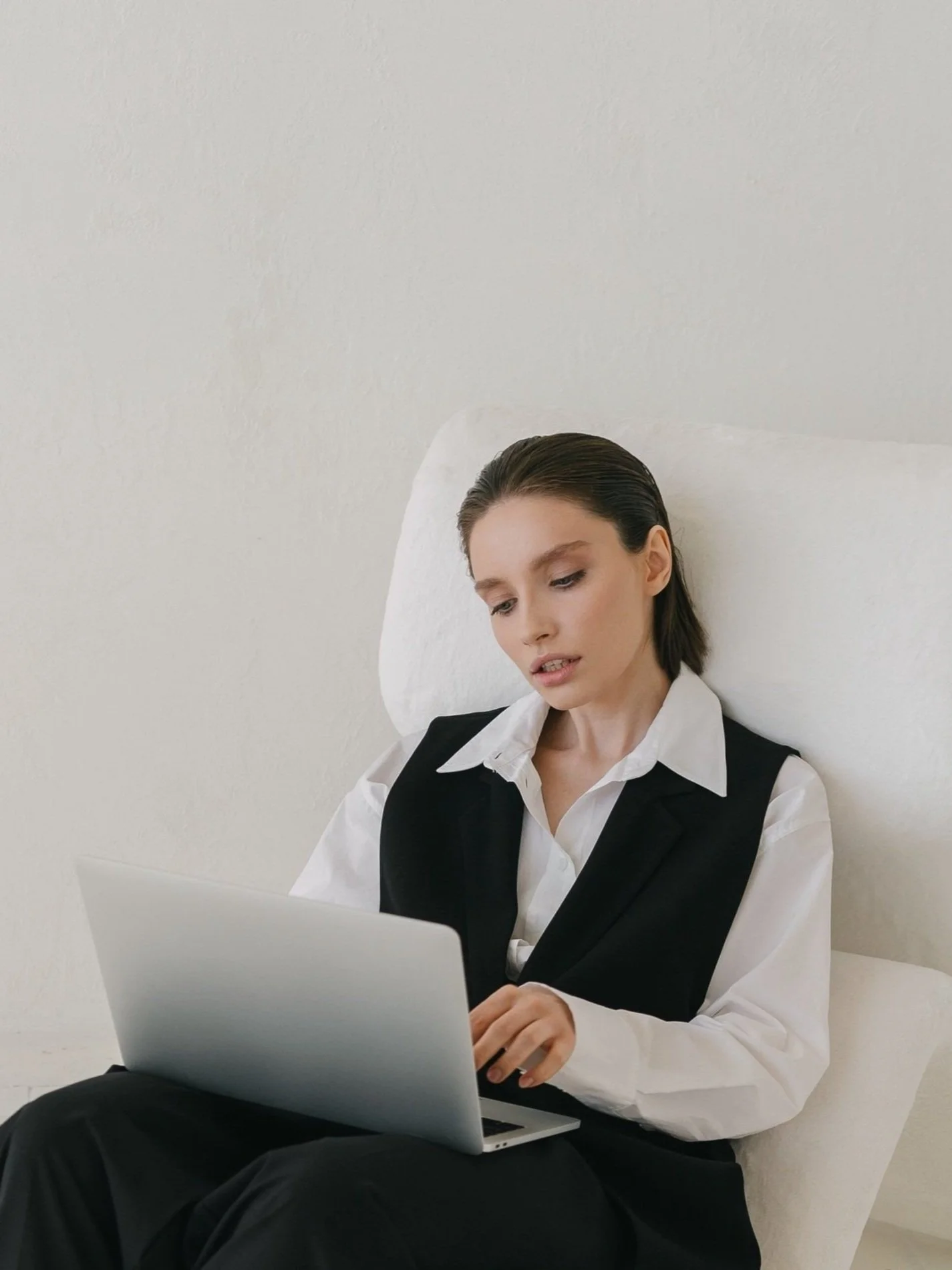 A young woman with short dark hair sitting on a white pillow against a white wall, looking at her laptop.