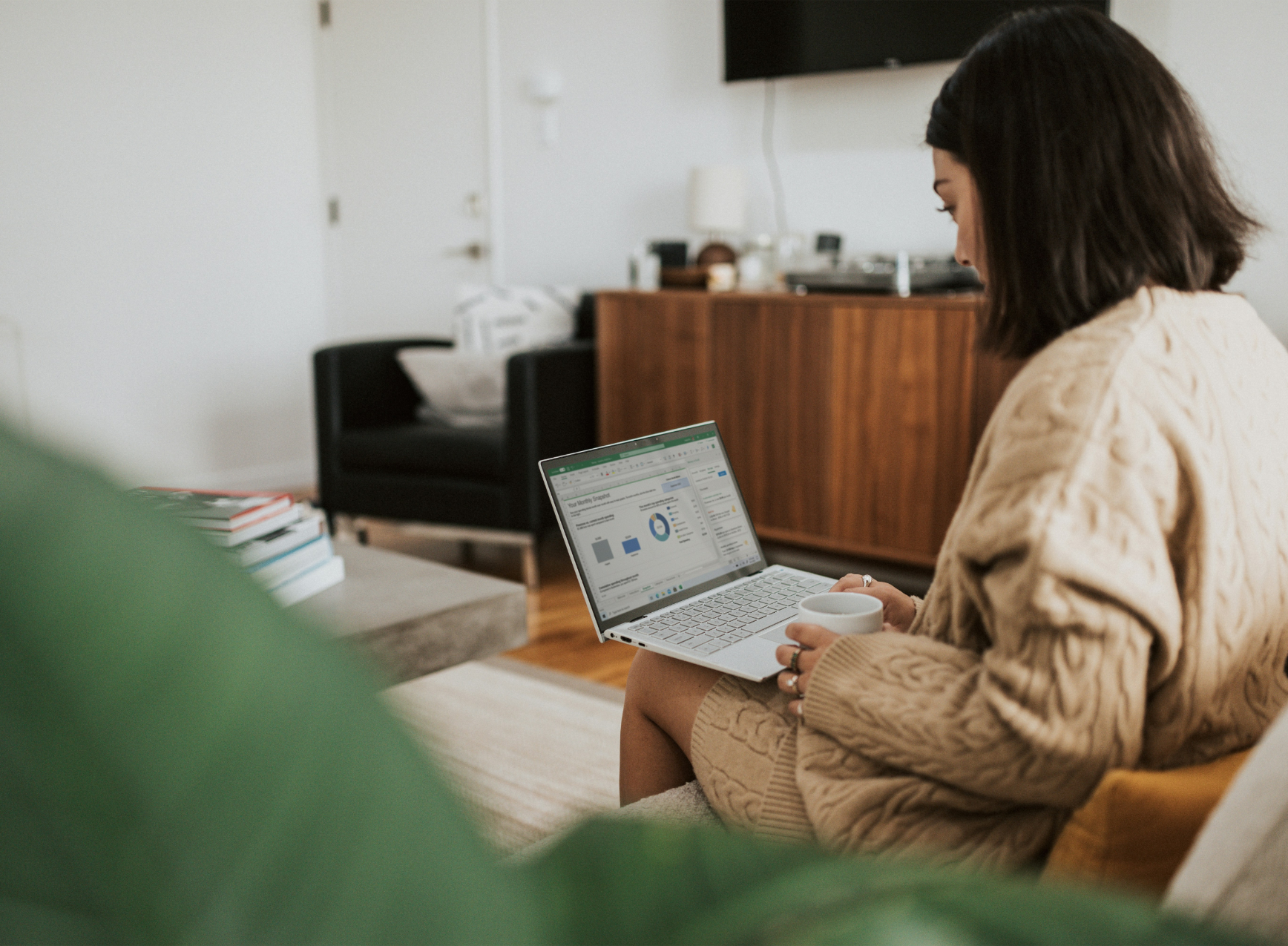 A woman sitting on a sofa in a living room, working on a laptop that displays a spreadsheet or data analysis dashboard. She is holding a white mug in her right hand and wearing a beige sweater. In the background, there is a wooden sideboard, a flat-screen TV mounted on the wall, and some decorative items.