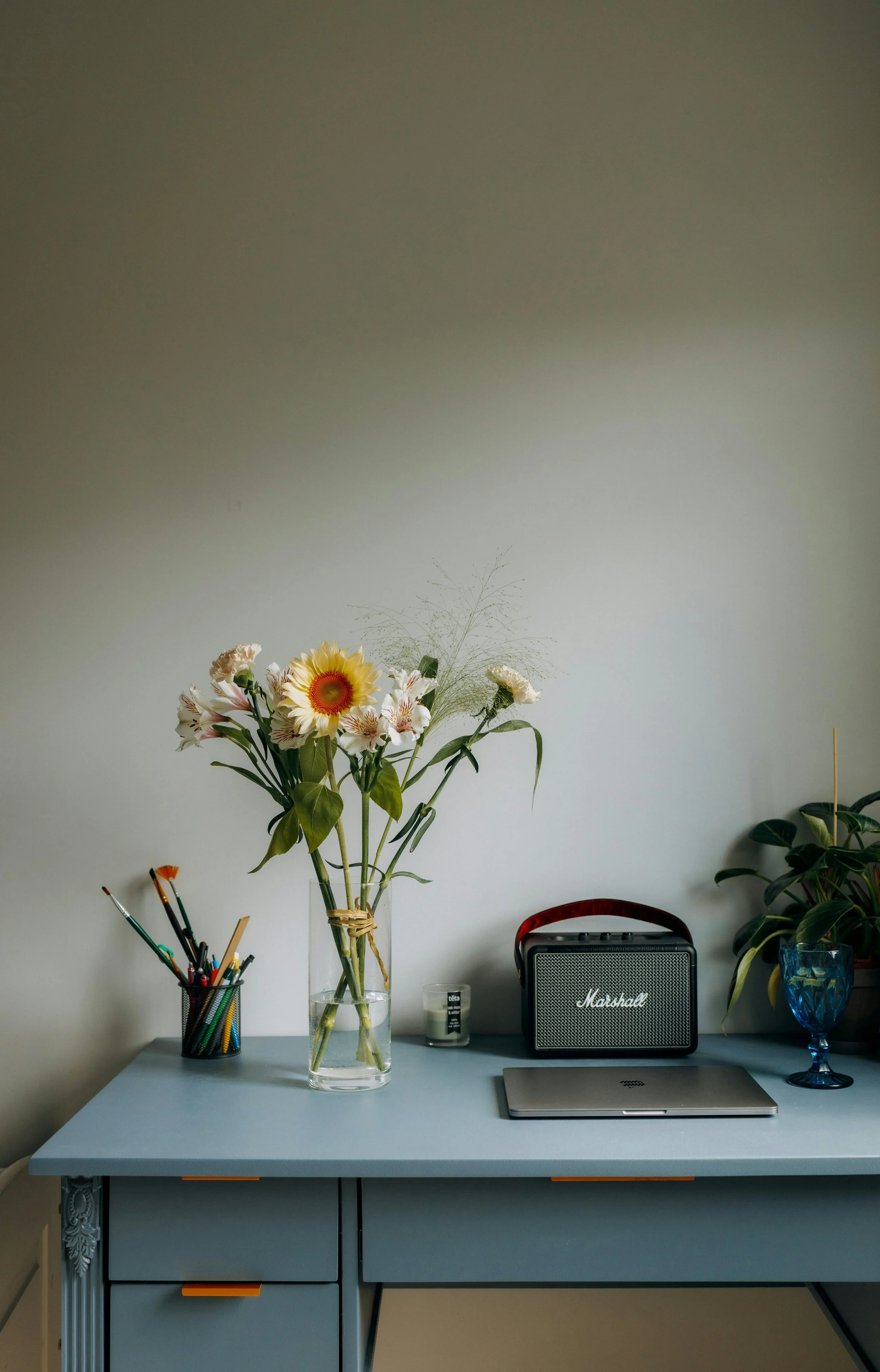 A desk with a vase of flowers, a container of colored pencils, a candle, a laptop, a Marshall speaker, and a potted plant against a plain white wall.