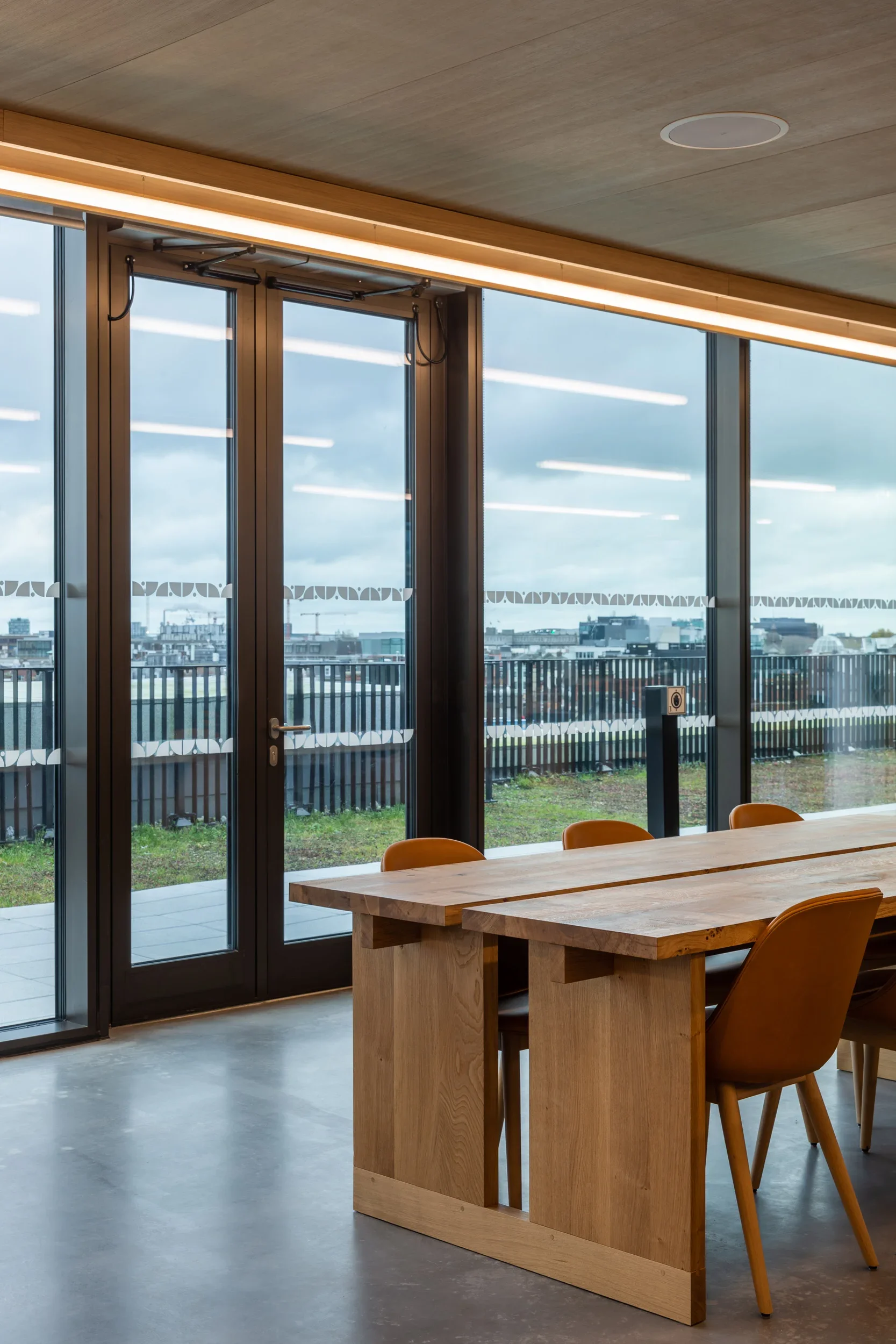 Interior of a modern room with large glass windows. There is a wooden table and chairs, with a city skyline visible in the background.
