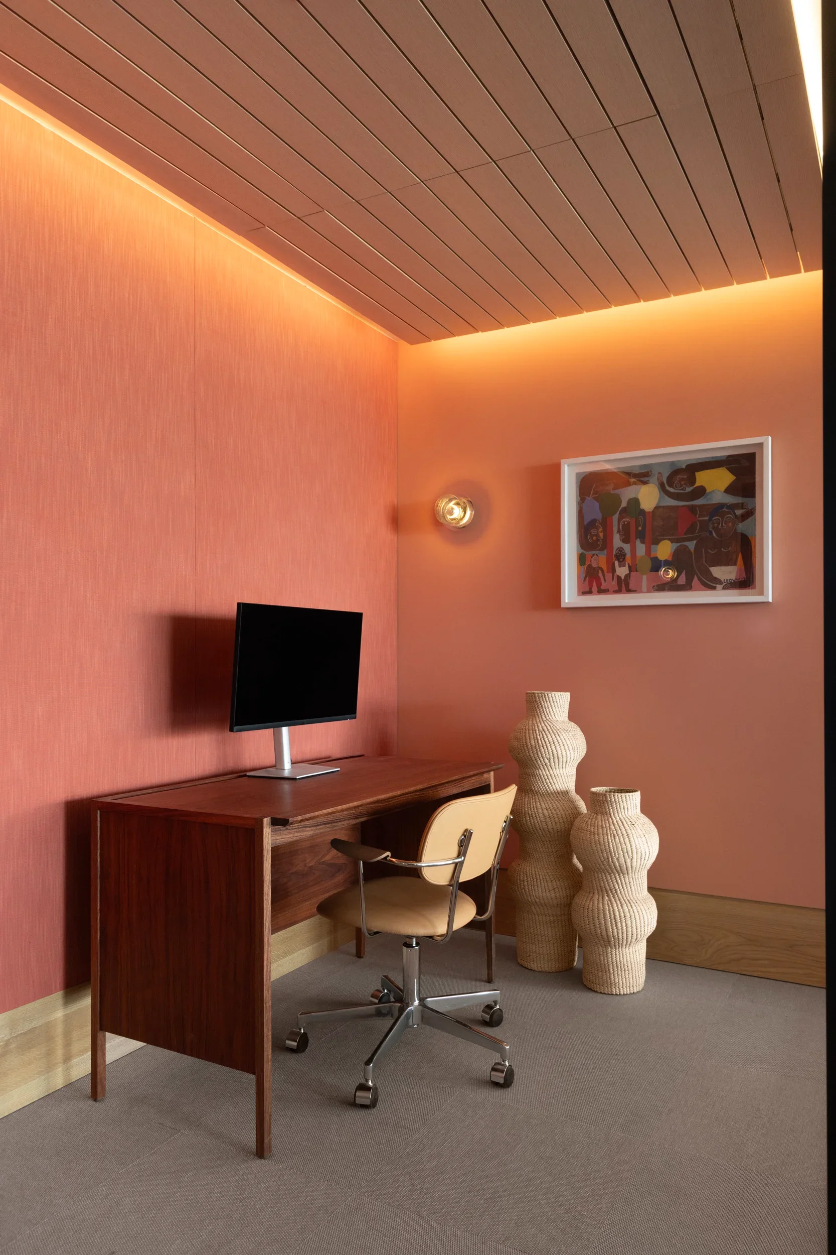 Modern home office with a warm color palette. Features a wooden desk with a flat-screen monitor and beige office chair. Decorative woven vases and a framed colorful artwork hang on the pinkish wall. The ceiling is wood-paneled with warm LED lighting.