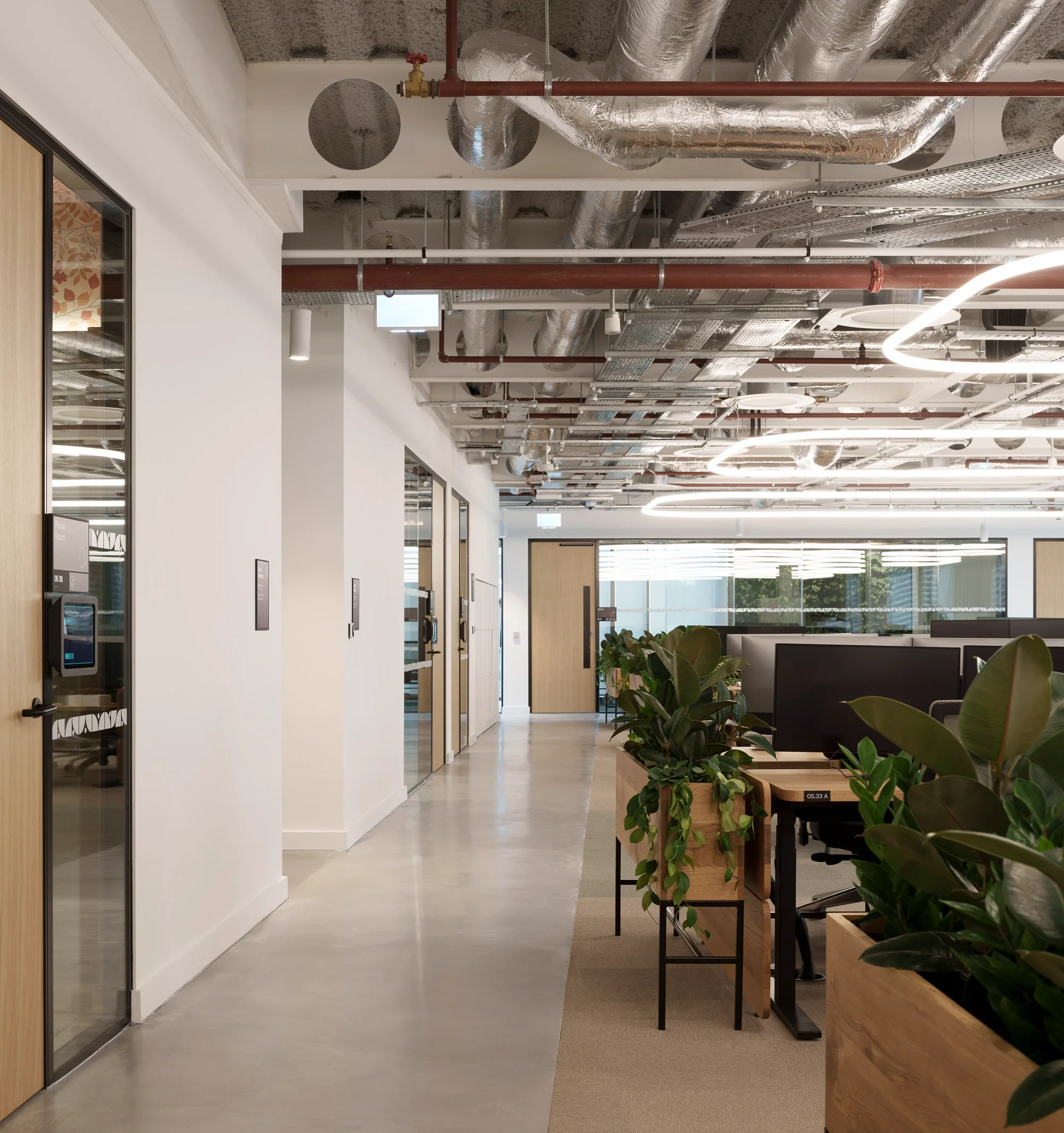 Modern office space with plants and workstations, featuring exposed ductwork and ceiling fixtures.