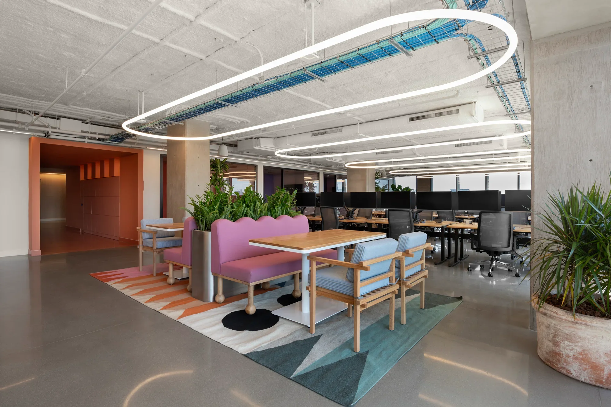 Modern coworking space with a casual lounge area featuring pink and blue armchairs, wooden tables, and potted plants, with workstations and office chairs in the background, illuminated by stylish circular ceiling lights.