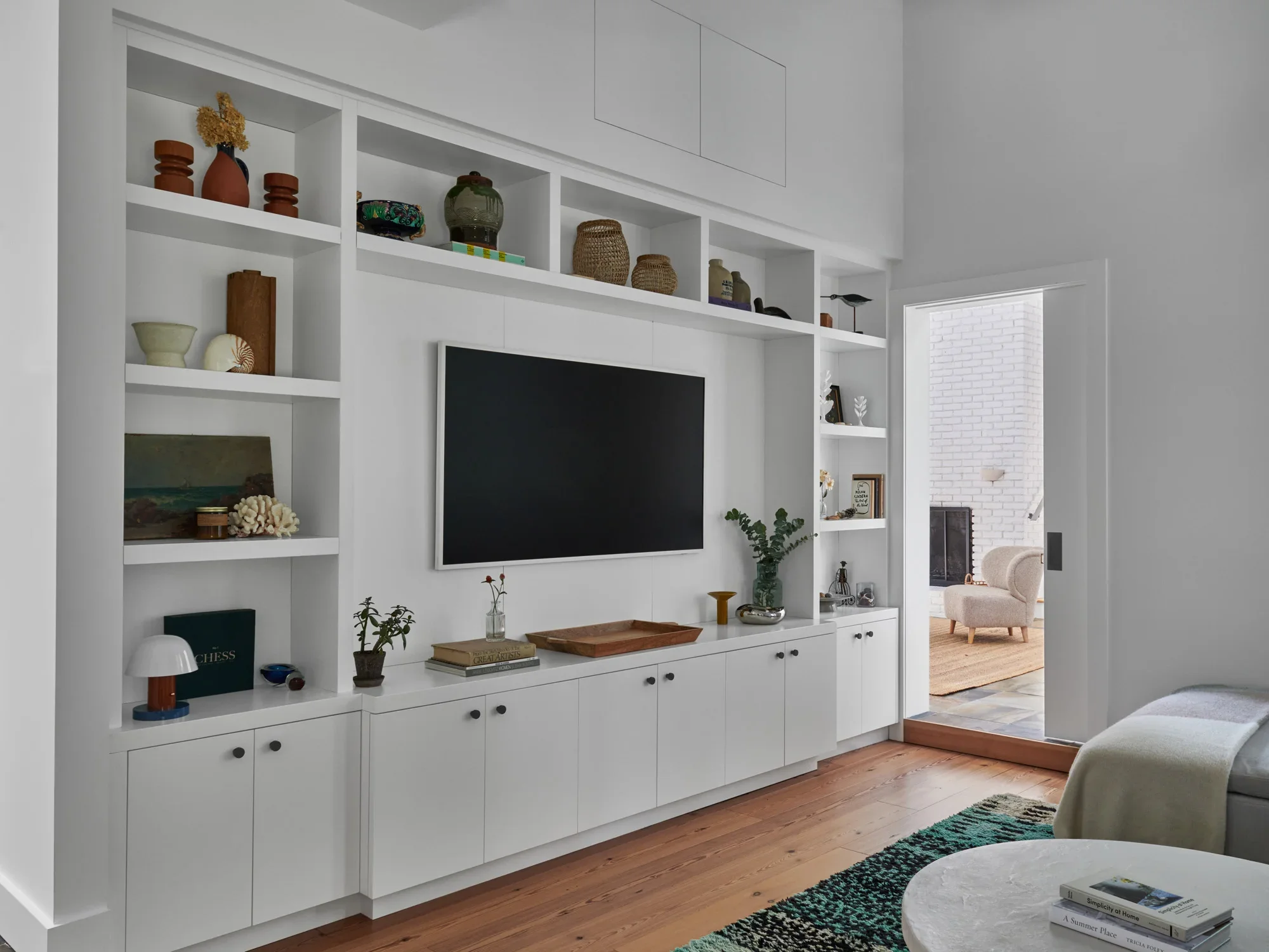 Living room with a white built-in shelf holding vases, books, and artwork. A flat-screen TV is mounted on the shelf. There is a doorway to another room with white brick walls, a beige armchair, and a natural fiber rug.