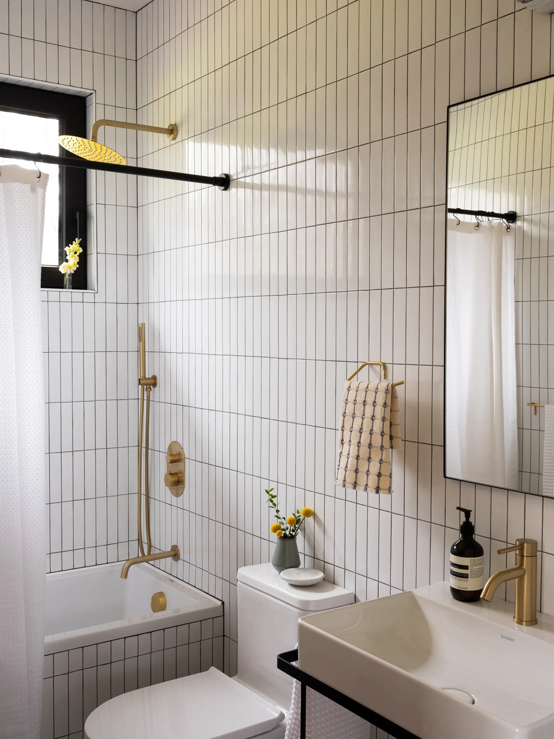 Modern bathroom with white tiled walls, black window frame, gold and black fixtures, a shower with a yellow rain showerhead, a white bathtub, a small white vanity sink with a gold faucet, a mirror, a black soap dispenser, a towel rack with a checkered towel, and a white toilet with a small vase of yellow flowers on top.