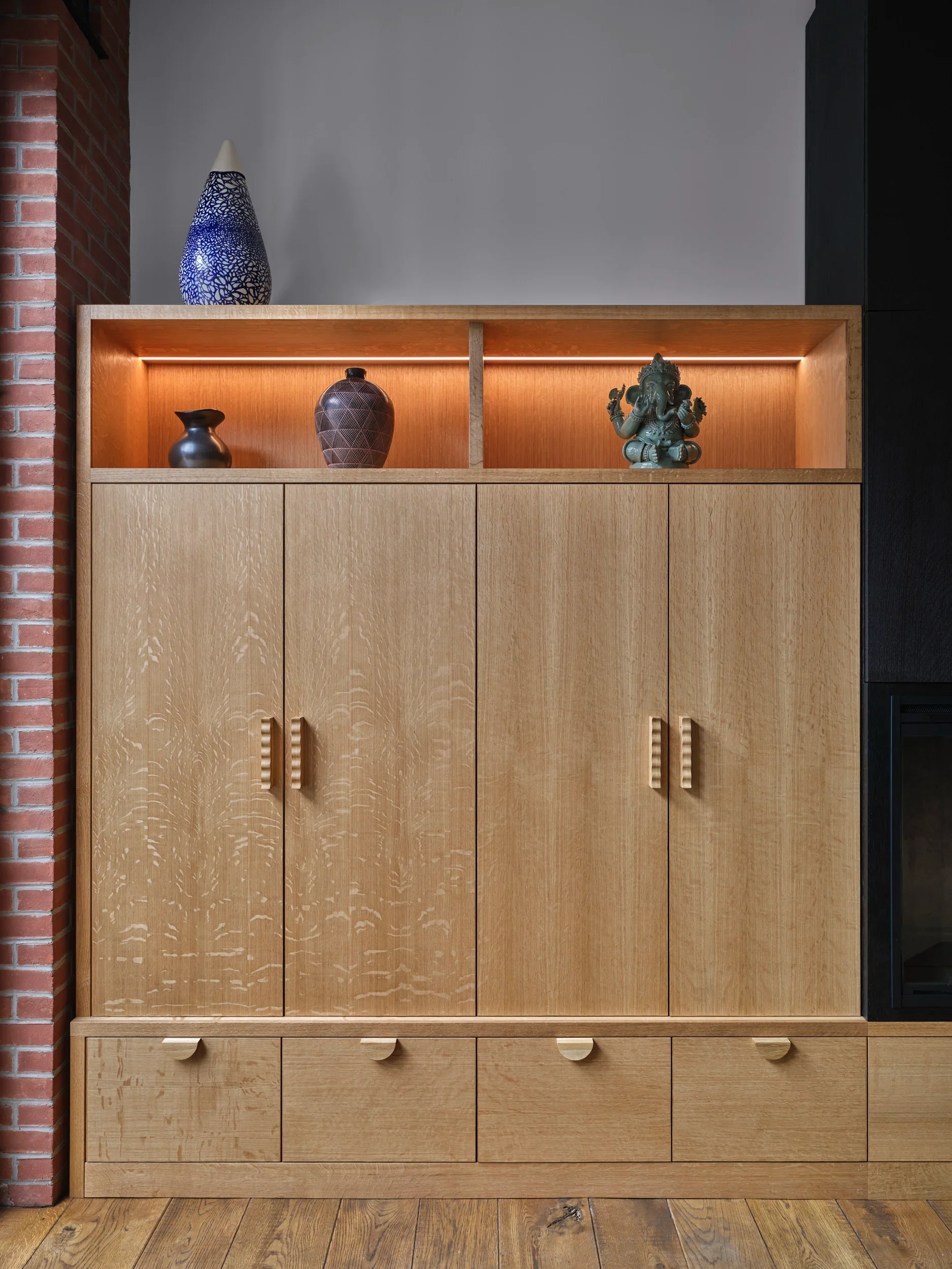 Light wooden cabinet with four doors and four drawers, decorative vases and a sculpture of Lord Ganesha on the top shelf, against a plain wall with brick on the side.