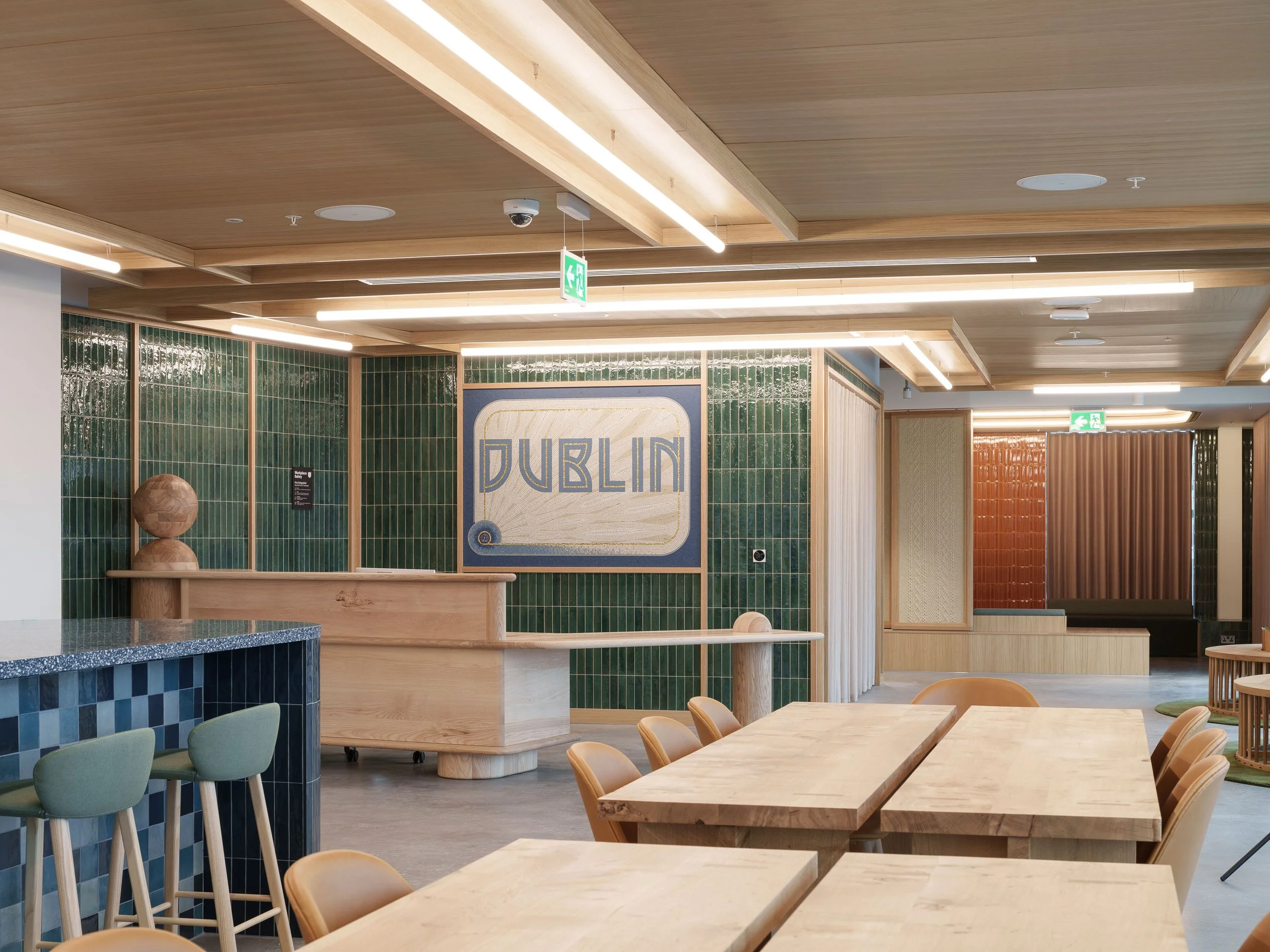 Interior of a modern dining area with wooden tables and green chairs, featuring green tiled walls and wooden ceiling panels with lighting fixtures. A sign with the word "DUBLIN" is visible on the wall.