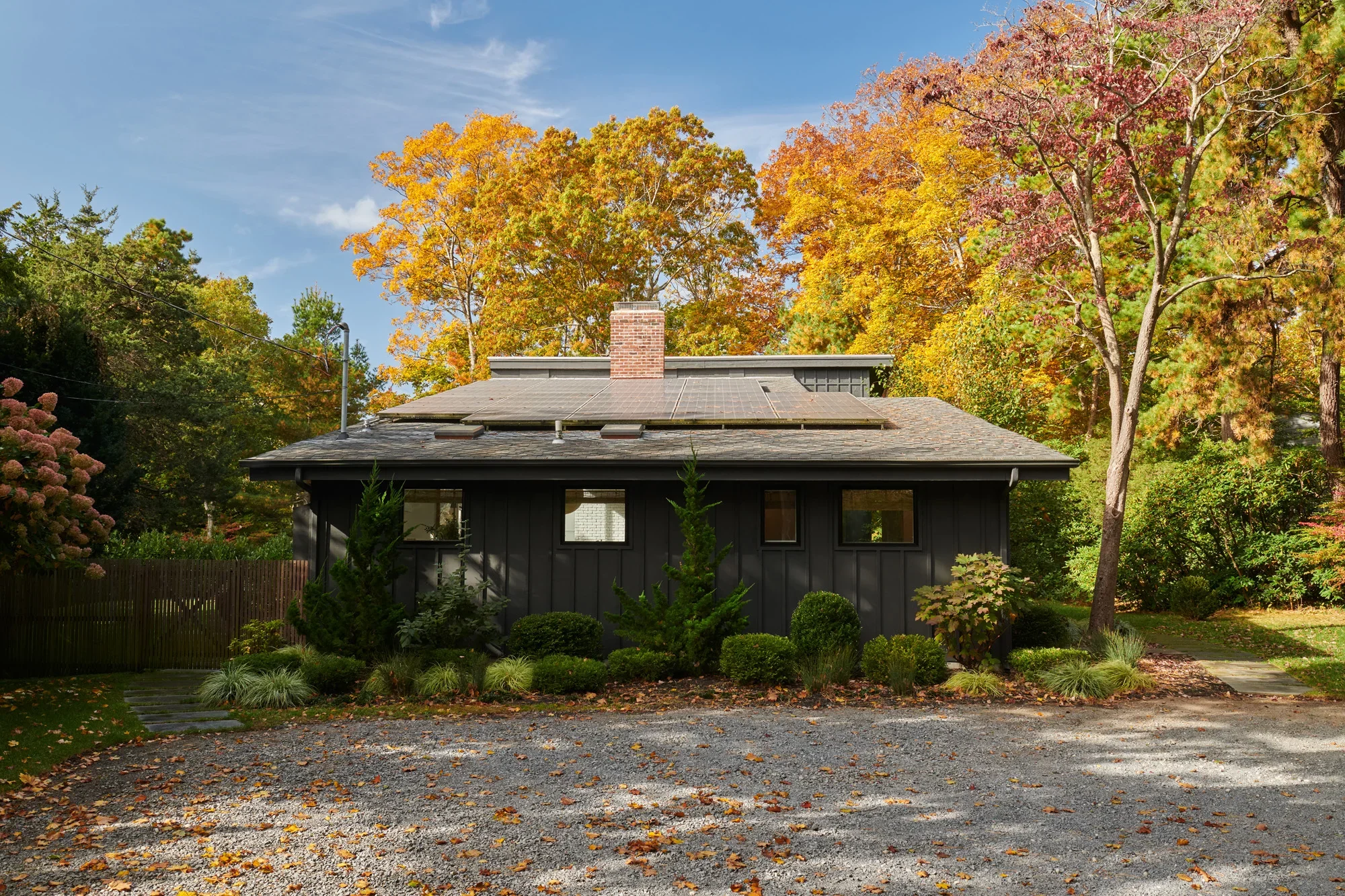 A house with a dark exterior and a roof fitted with solar panels, surrounded by trees with autumn-colored leaves, a gravel driveway, and a small stone pathway.
