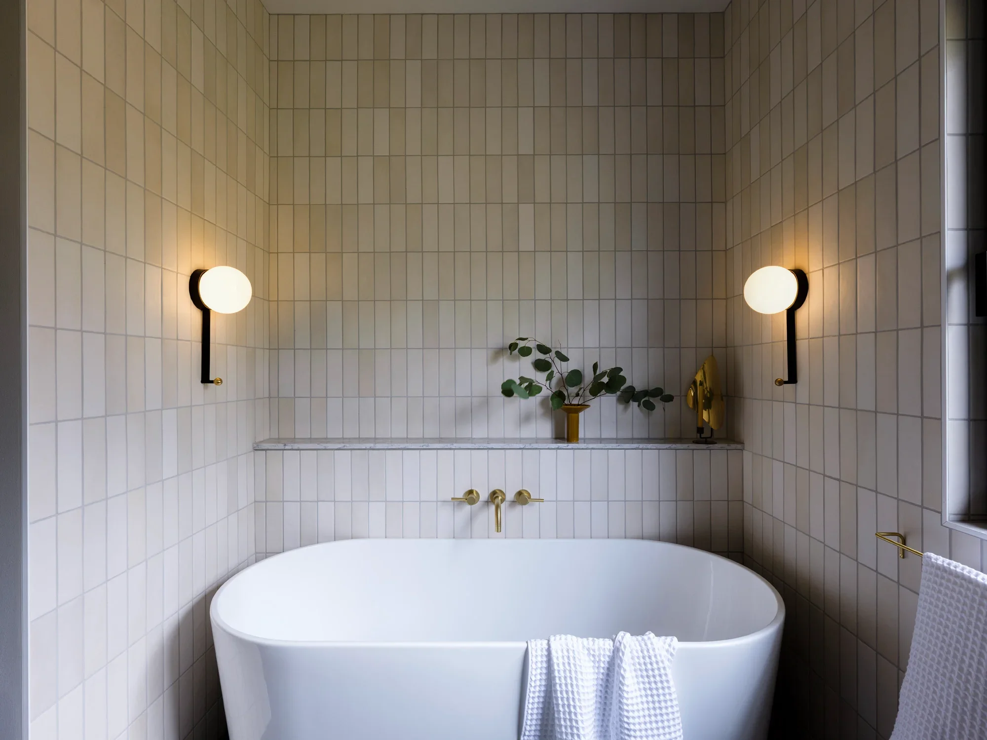 Modern bathroom with a freestanding bathtub, beige tiled walls, and black wall-mounted lamps. There's a small table with a vase and plant, and a window on the right letting in natural light.