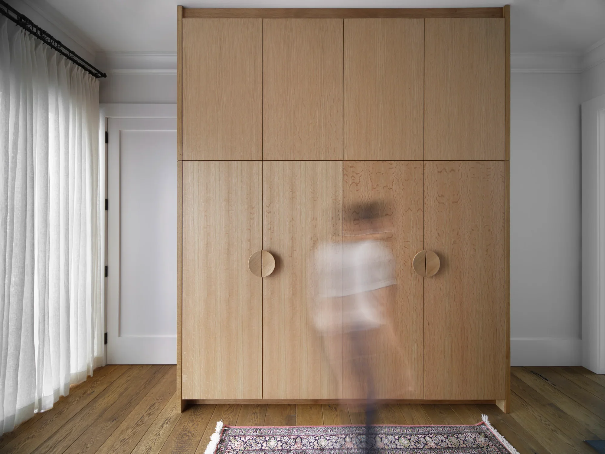 A large wooden wardrobe with six doors is placed against a wall in a room with hardwood flooring. A patterned rug is partially visible at the bottom of the image, and sheer white curtains hang on a window to the left.
