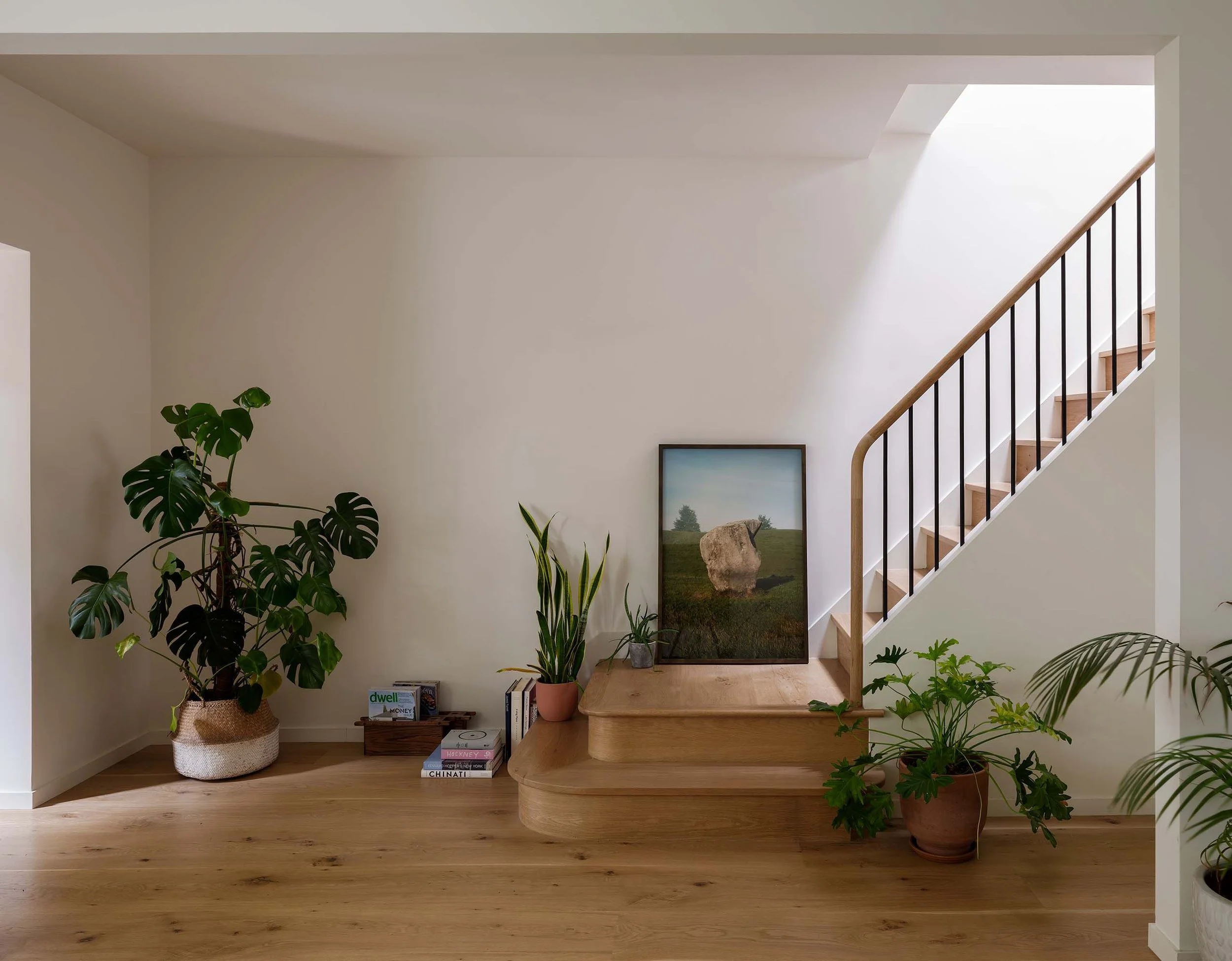 An interior view of a home staircase with wooden steps and black railing, surrounded by various potted green plants, a bookshelf with books, and wall art.