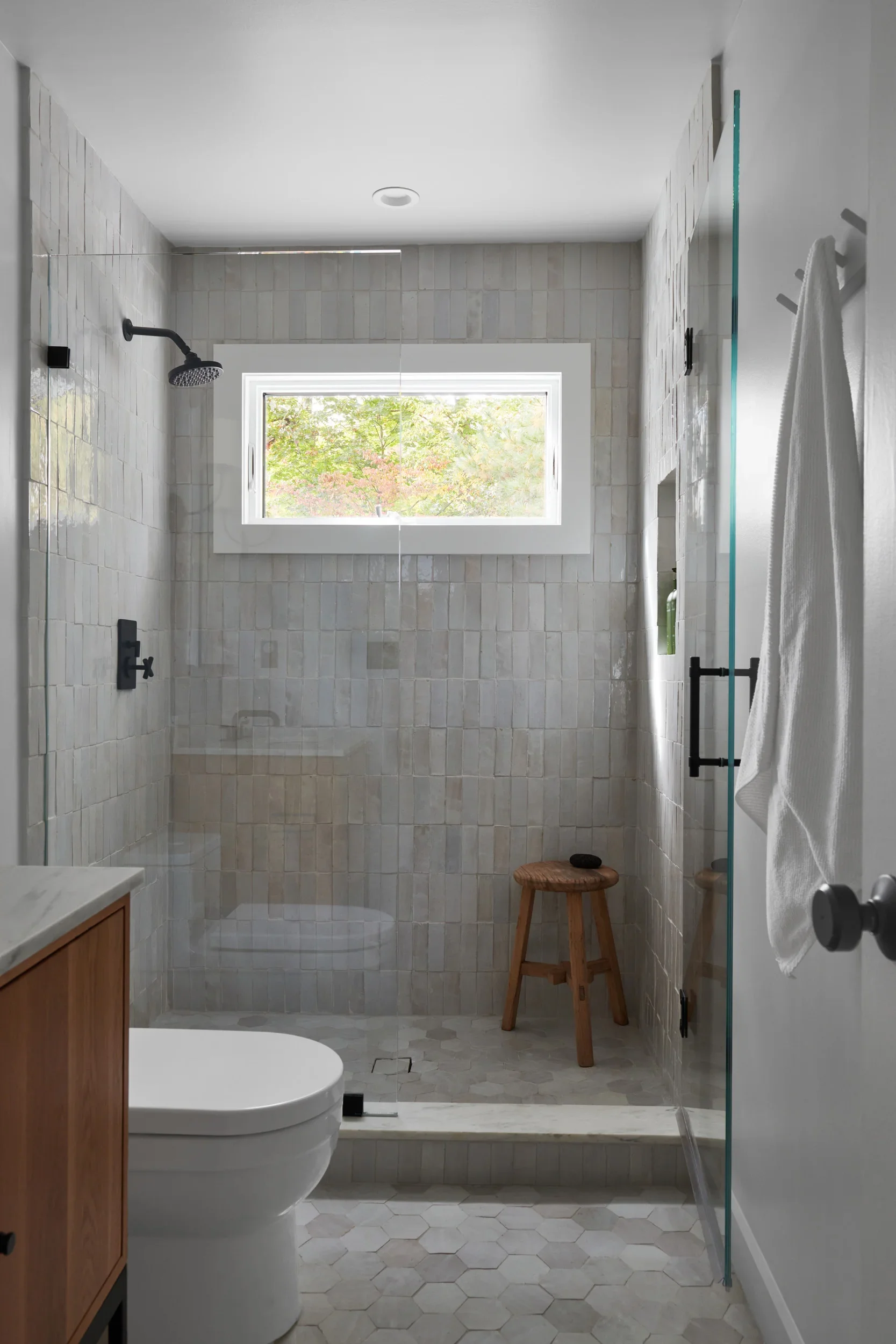 Modern bathroom with a walk-in shower featuring beige tile walls and a glass door. There is a window above the shower, allowing natural light, with a view of trees outside. The bathroom has a white toilet, a wooden stool, and a towel hanging on a rack.