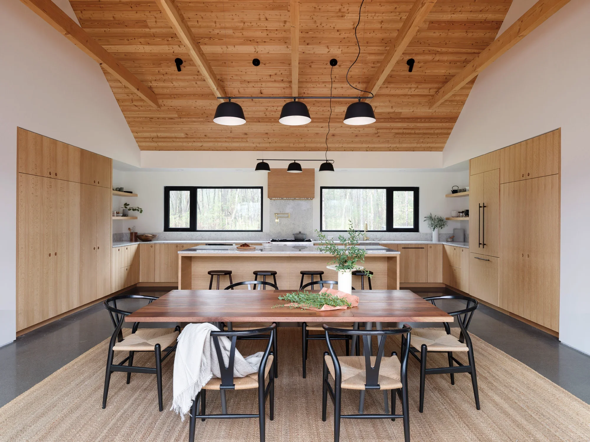 Modern kitchen with wooden cabinets, a wooden ceiling, black pendant lights, large windows, and a dining table with black chairs.