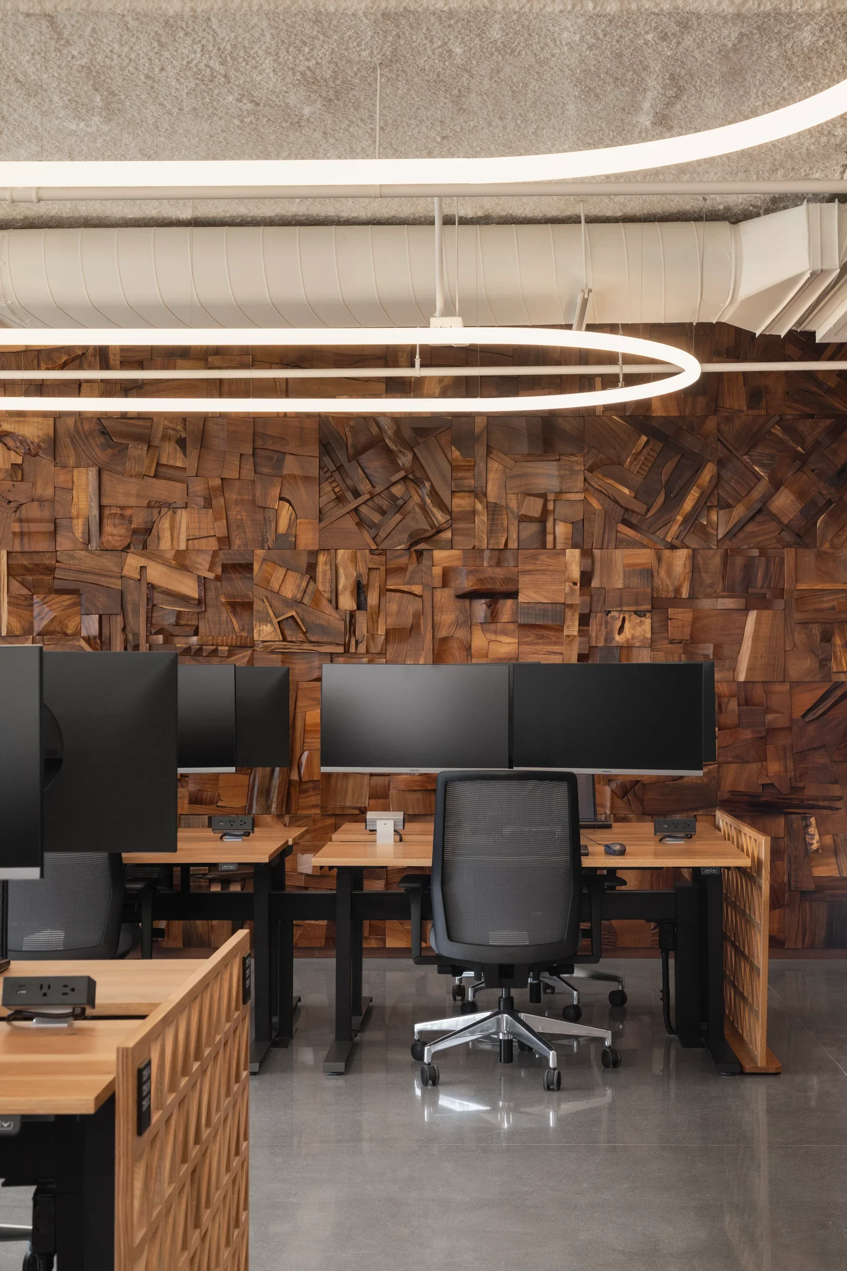 Modern office workspace with multiple desks, black computer monitors, ergonomic chairs, and a decorative wooden accent wall. Overhead lighting includes circular fixtures and an exposed duct ceiling design.