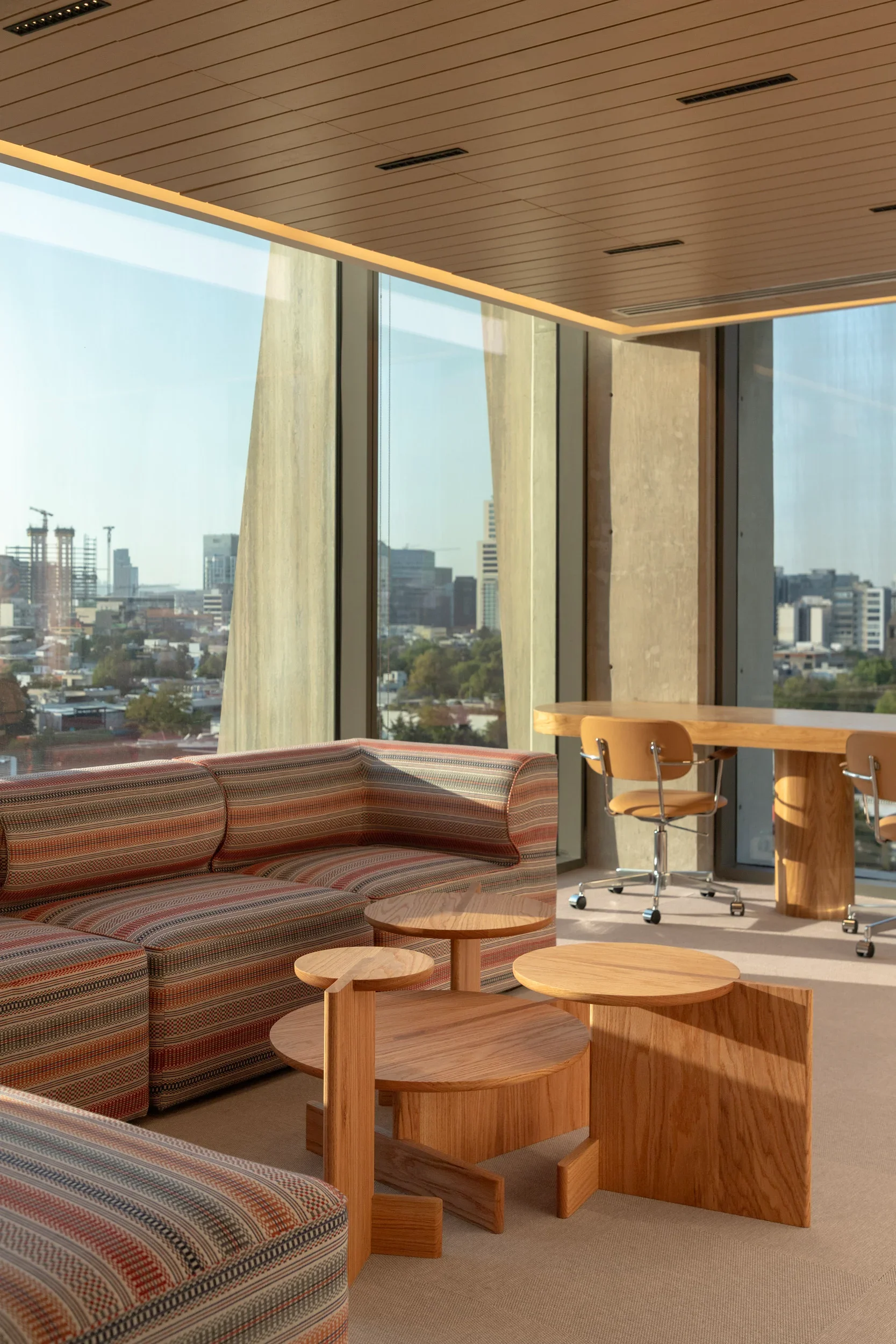 Modern office space with large glass windows, tan and wooden furniture including a striped sofa, round tables, and a desk with a yellow chair, overlooking a city skyline.