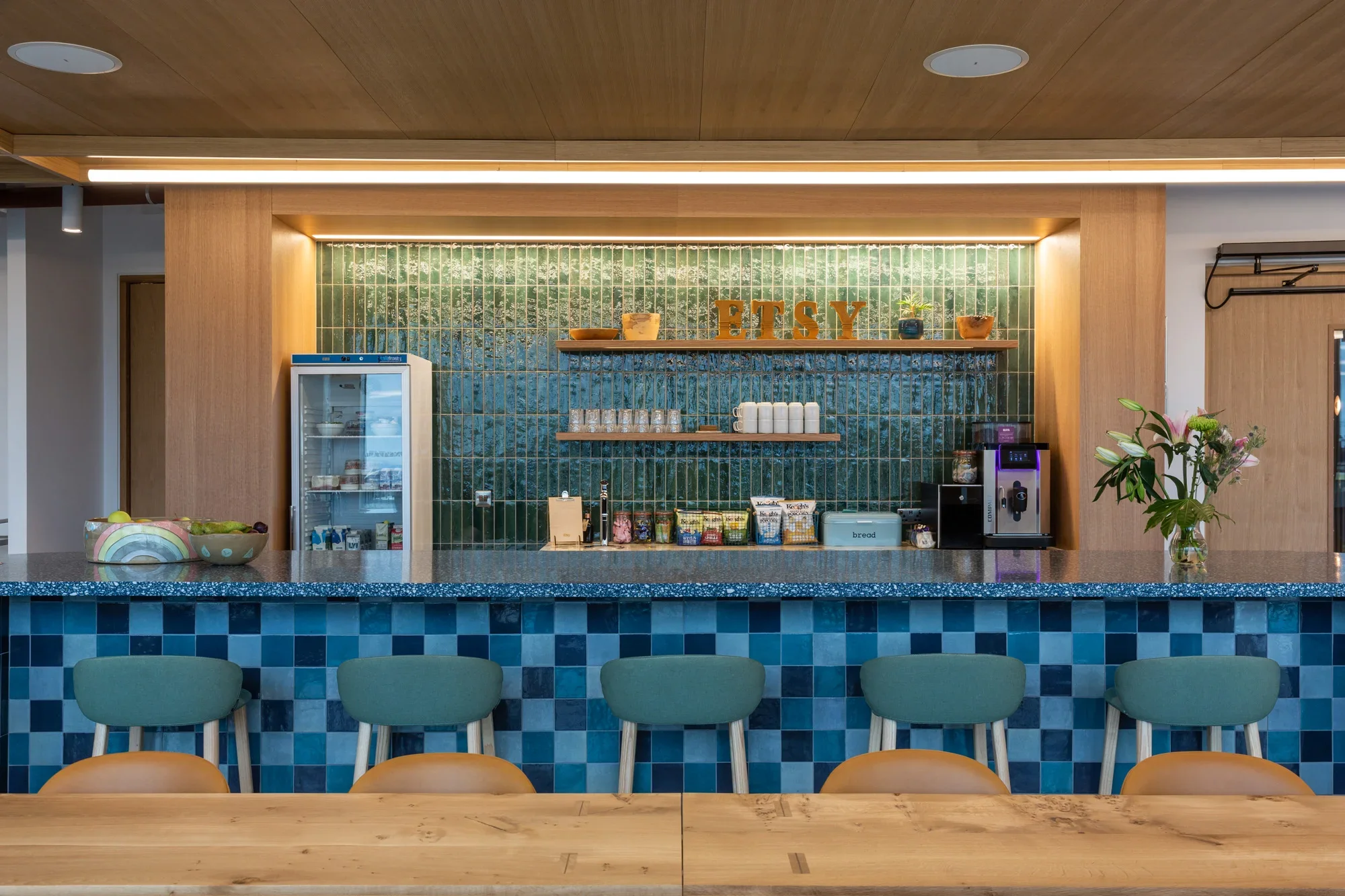 A modern cafe or breakfast bar with a blue tiled front, green chairs, and a wooden table in the foreground. The background features a green tiled wall with shelves holding bowls, mugs, and decorative items, along with a fridge and coffee machine.