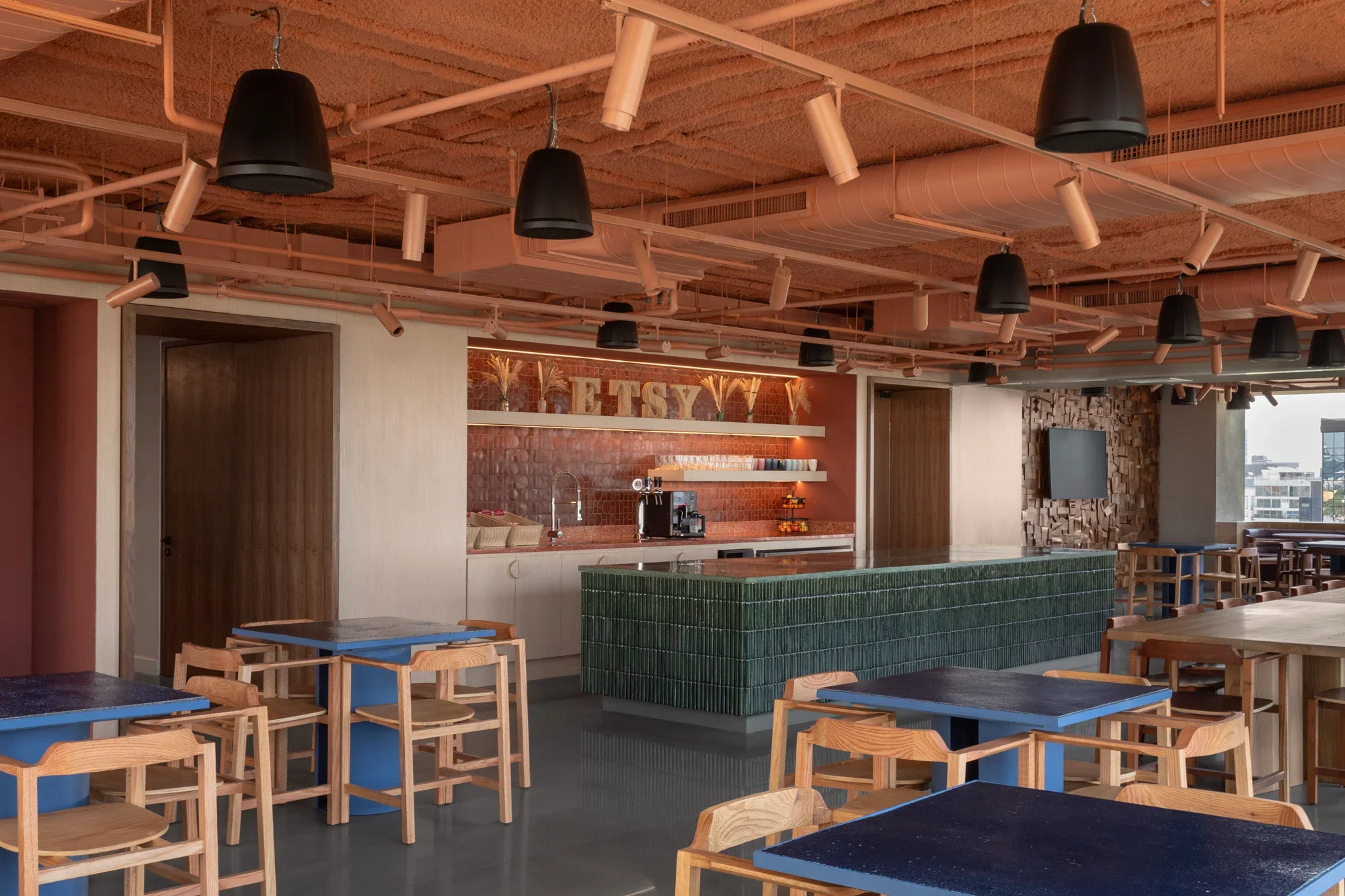Modern cafe interior with wooden chairs and tables, black pendant lights, a green tiled counter, and a brick wall with the word "TSETY" displayed.