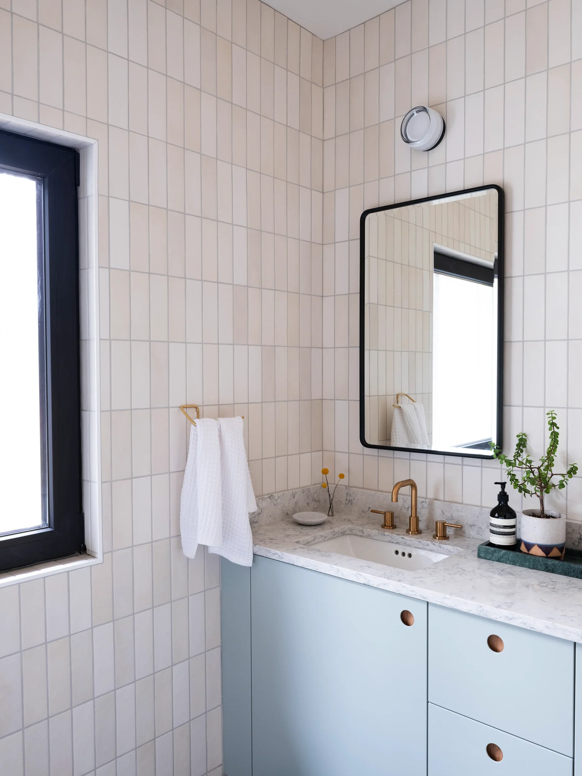 Bathroom sink area with a marble countertop, a gold faucet, and gold handles on the cabinet. There is a mirror on the tiled wall, a window with a black frame to the left, and a small potted plant on the counter. White towels hang on a gold towel rack.