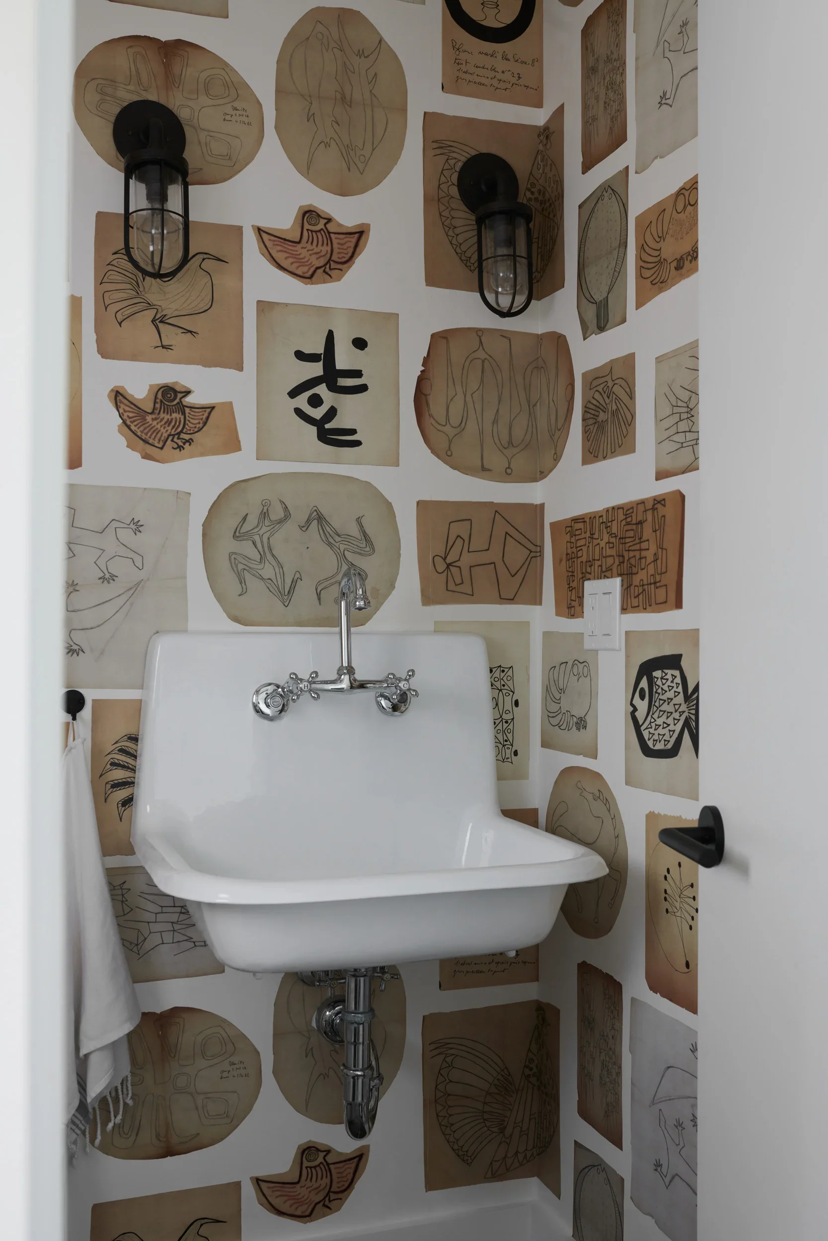 A white bathtub with a chrome faucet, installed in a corner with walls decorated with various abstract and animal sketches on brown paper. Two black cage-like wall lights are mounted above the bathtub, illuminating the art on the walls.