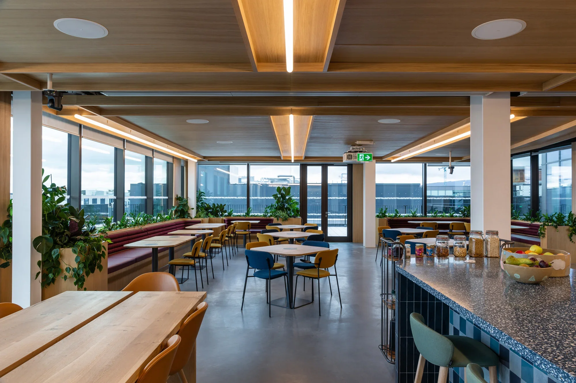 Modern dining area with wooden ceiling panels and large windows, featuring tables and chairs, and a breakfast bar with jars of cereal and fresh fruit.