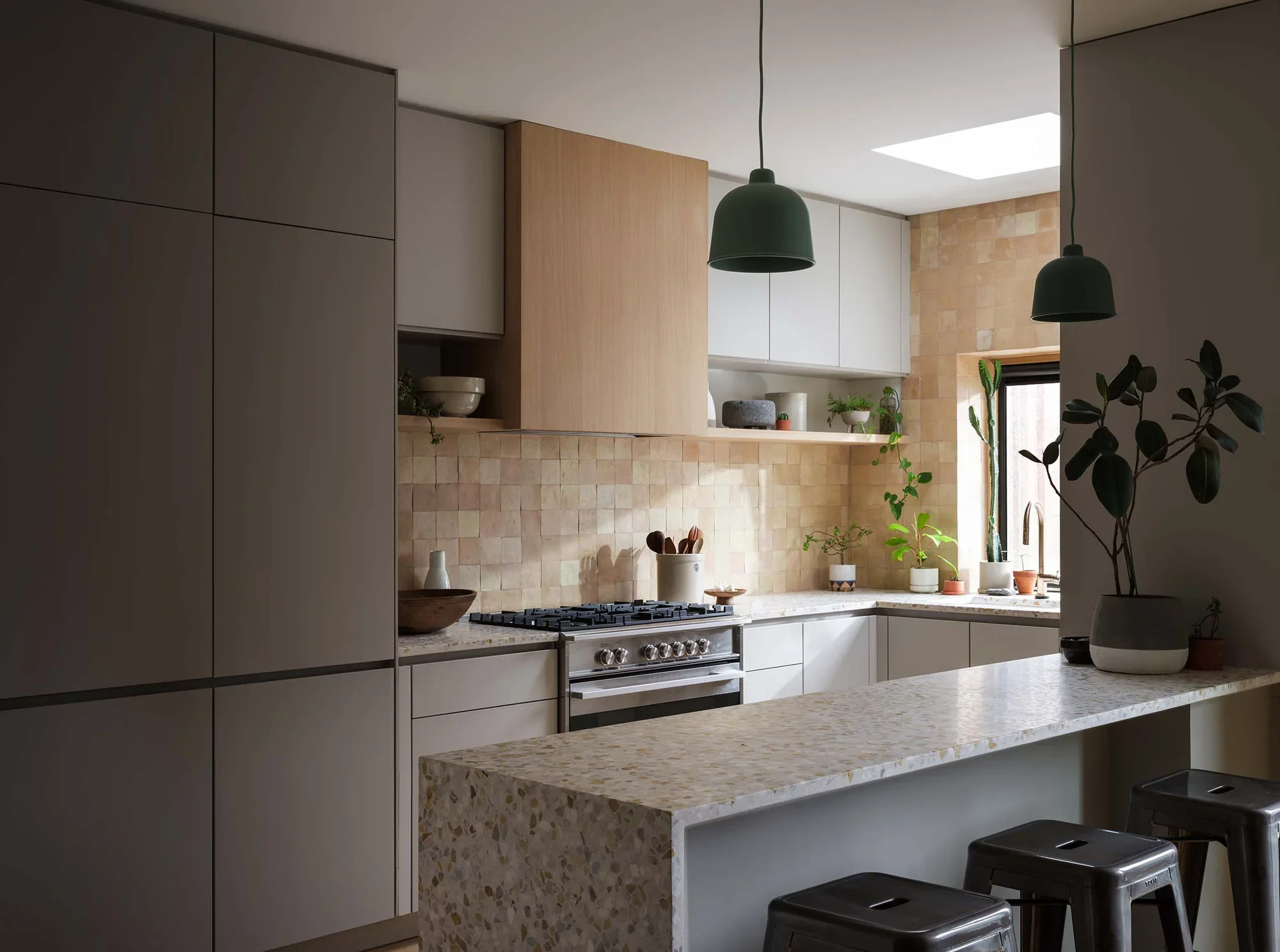 Modern kitchen with white and wooden cabinets, a speckled marble countertop island, and various potted plants on the window sill. Two black stools are at the island, and two green pendant lights hang from the ceiling.