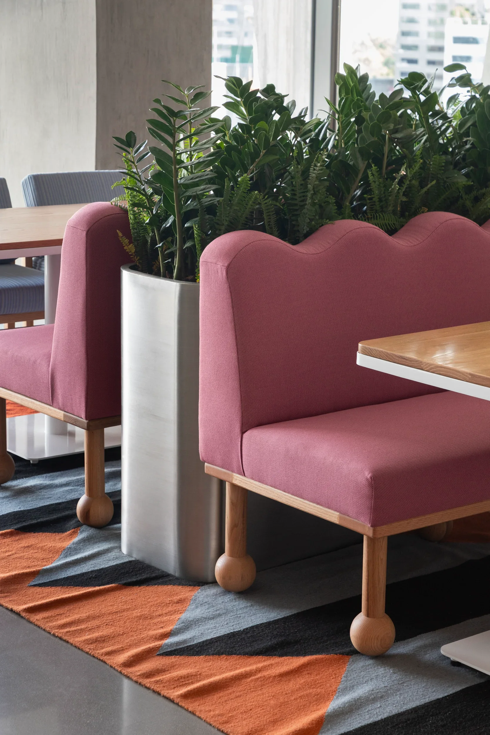 Interior space with pink upholstered chairs, a large plant in a white planter, a colorful striped rug, and windows showing a cityscape.