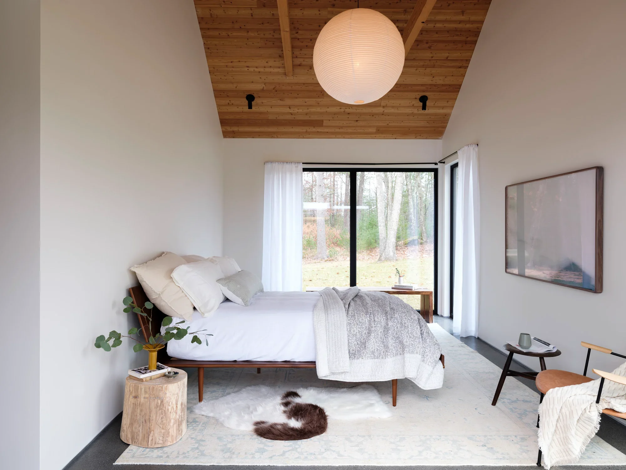 A modern bedroom with a wooden ceiling, white walls, and a large window showing trees outside. The bed has white bedding and pillows, with a potted plant on a small side table, and a furry rug on the floor.