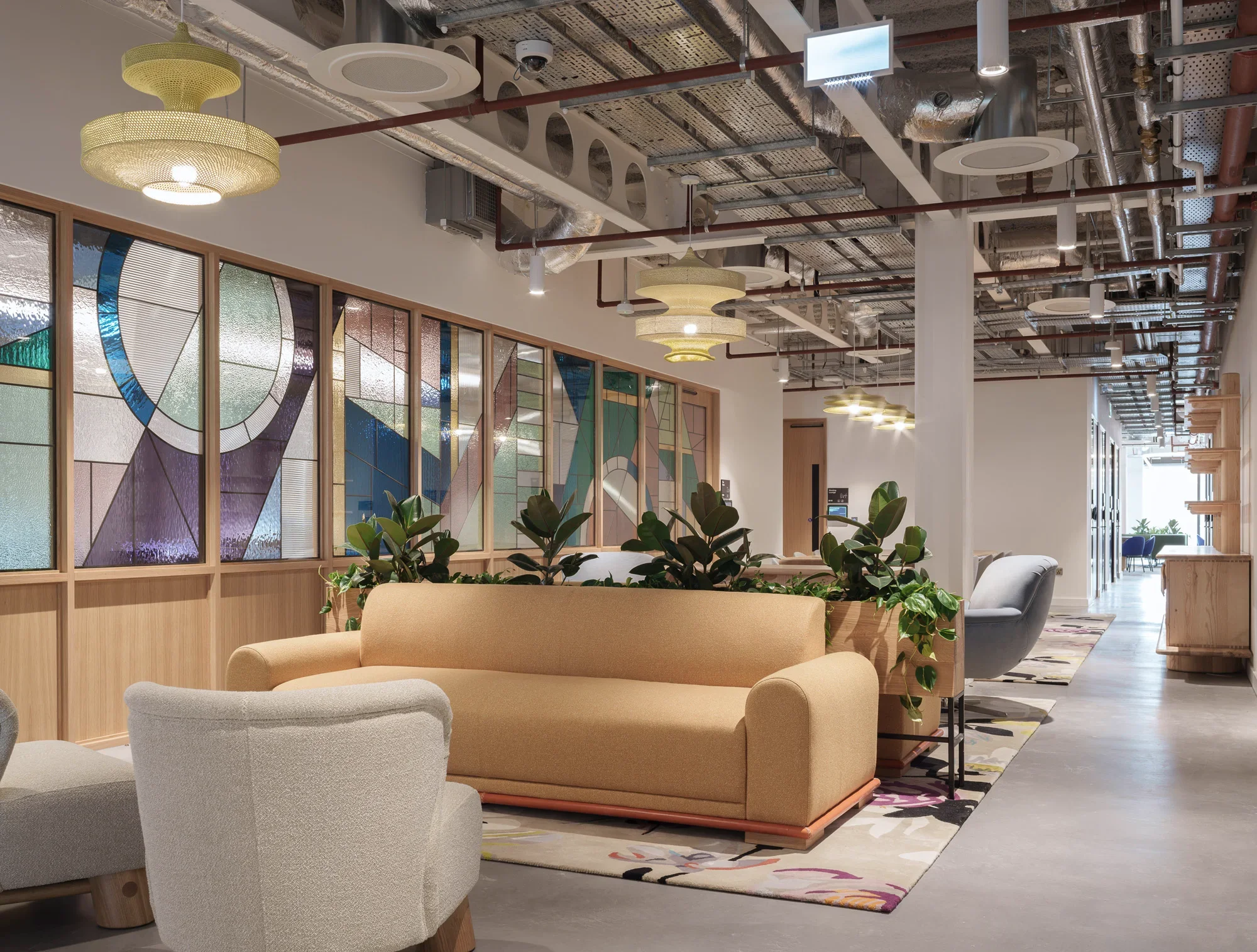 Modern office or co-working space with beige and gray upholstered seating, a large colorful geometric stained glass window, hanging light fixtures, an open ceiling with visible ductwork and lighting, and potted plants.