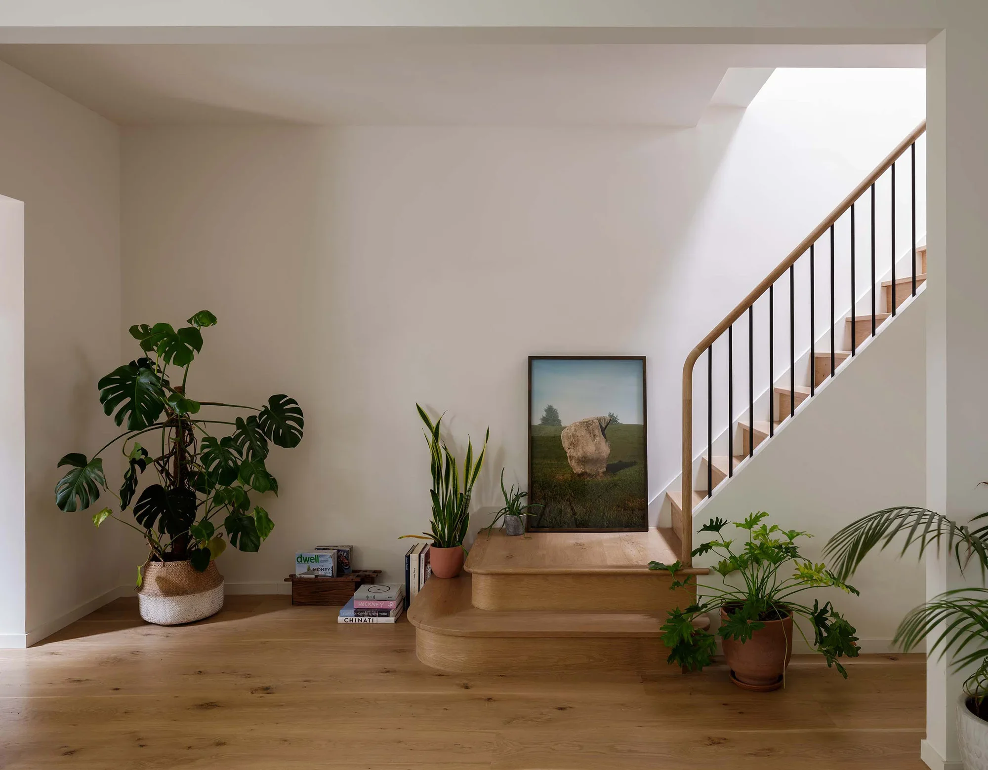 Interior view of a staircase landing decorated with various green houseplants, books, and artwork, with sunlight streaming in through an upper window.