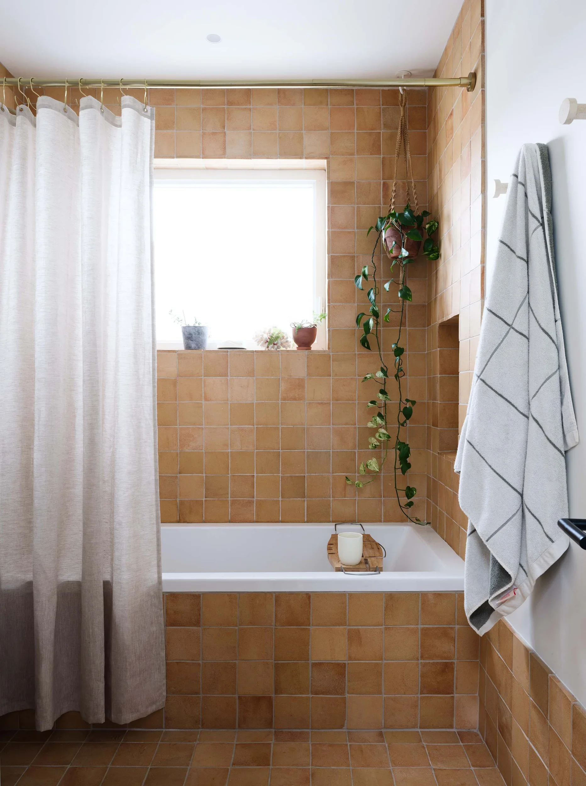 A bathroom with orange tile walls and floor. There is a bathtub with a wooden tray holding a white candle, a hanging potted plant with green leaves, a small window with potted plants on the sill, a white shower curtain on a gold rod, and a gray and white striped towel hanging on a hook.