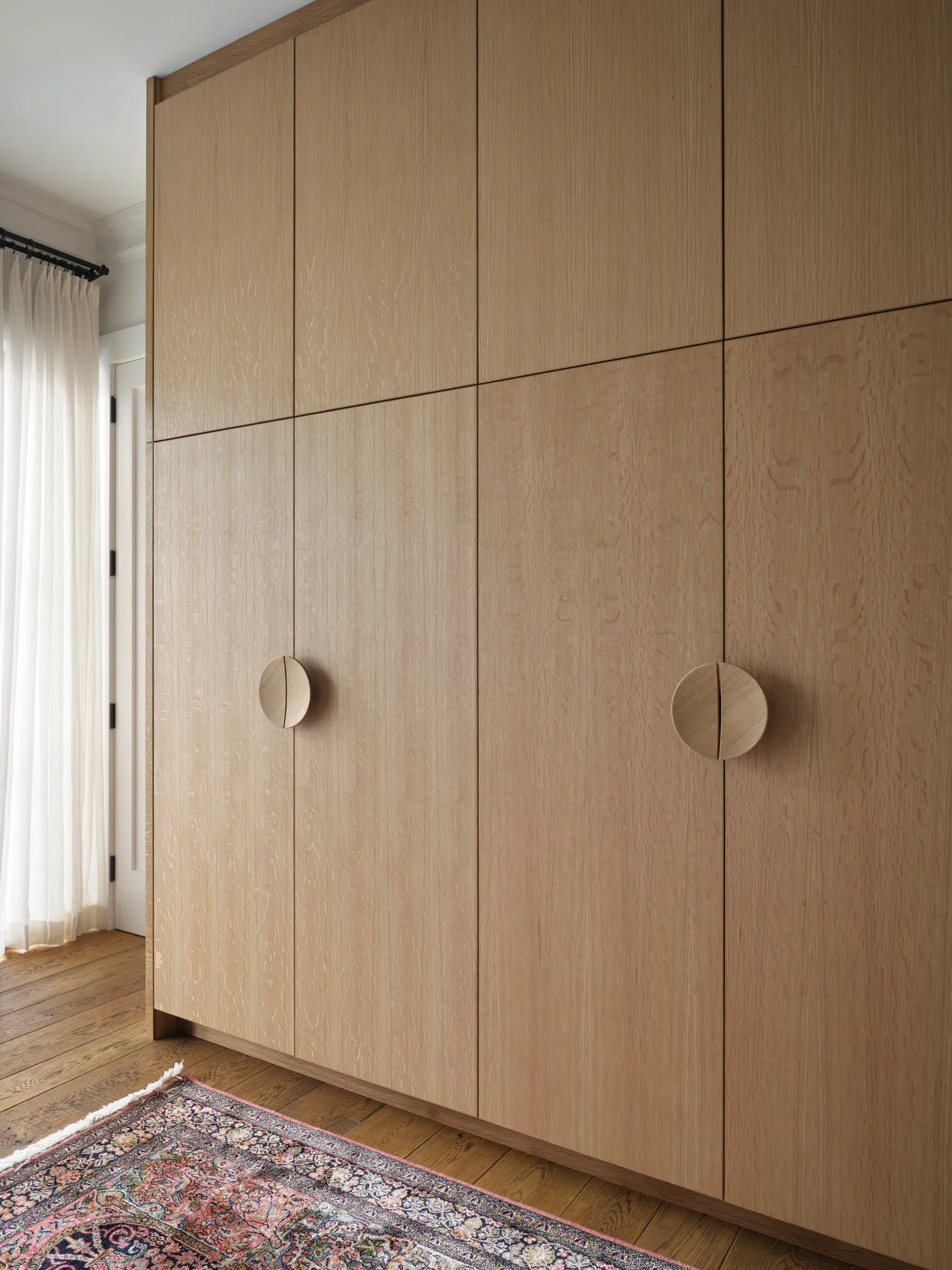 A large wooden wardrobe with circular handles in a room with a hardwood floor, a colorful rug, sheer curtains, and a door.