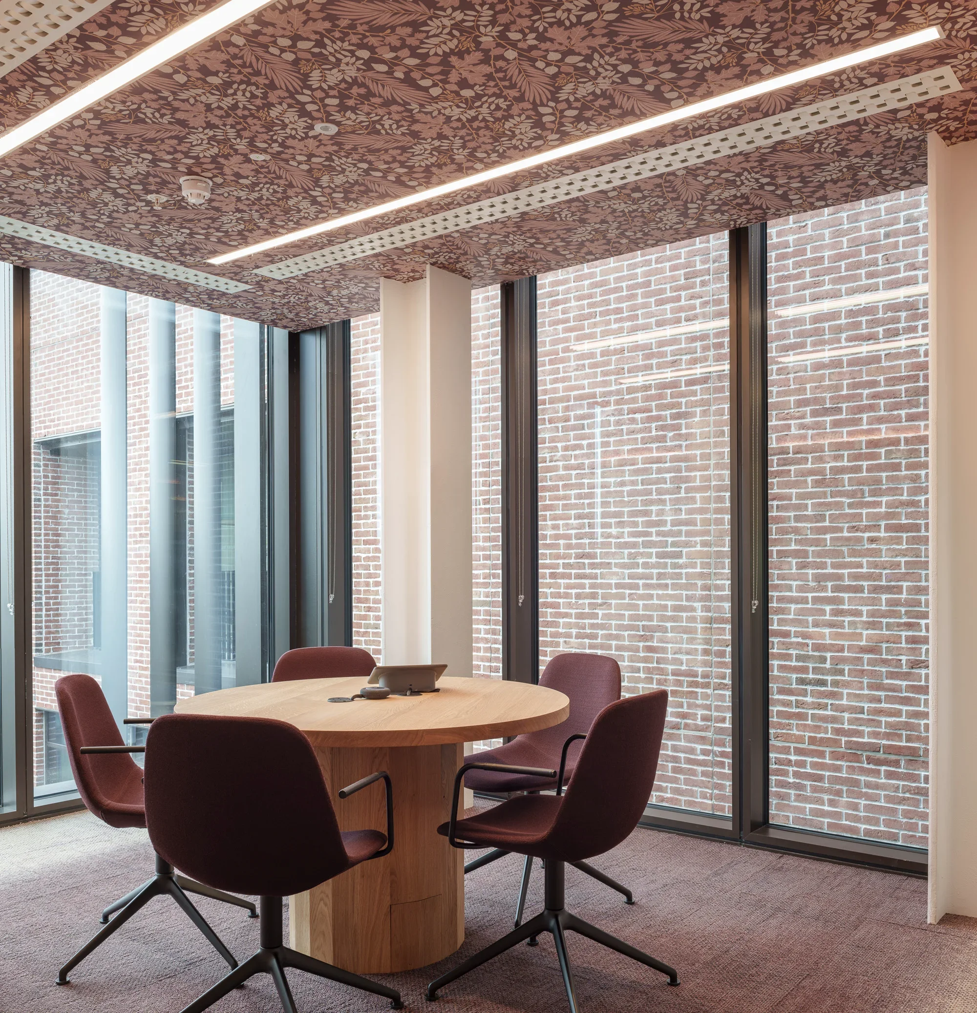 A conference room with a round wooden table surrounded by four red fabric office chairs. On the table, there is a landline telephone with a cord, some papers, and markers. The room has large floor-to-ceiling windows with black frames, revealing a brick wall outside. The ceiling has a floral patterned surface with purple and gold accents and features long, narrow LED light strips.