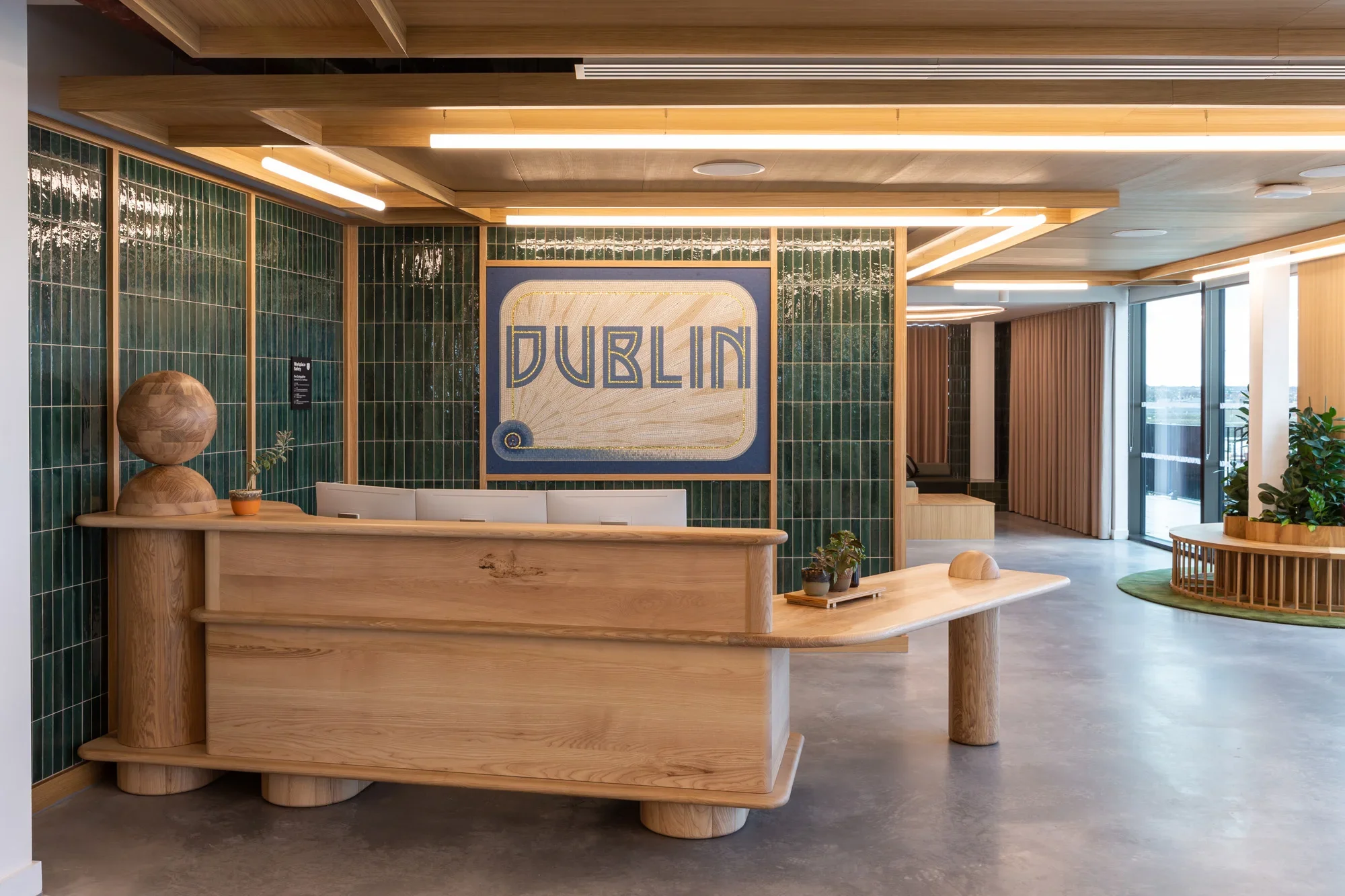 Modern lobby or reception area with wooden accents, green tiled walls, and a large sign reading 'DUBLIN' in front of a green tiled wall. There are decorative plants, a wood sculpture, and natural lighting from large windows.