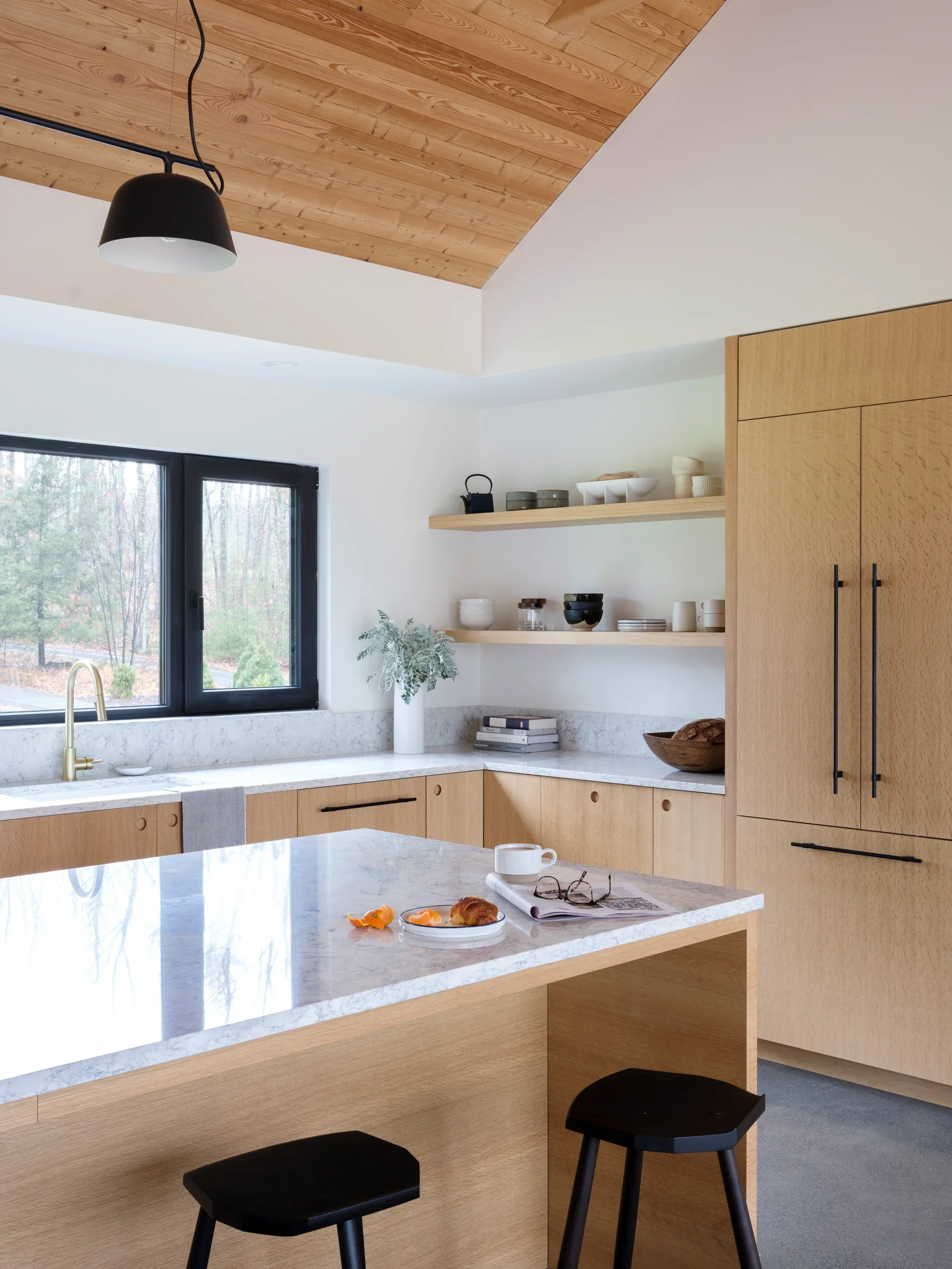 Modern kitchen with wooden cabinets, open shelves, a marble countertop, black stools, and a large window with a view of trees outside.