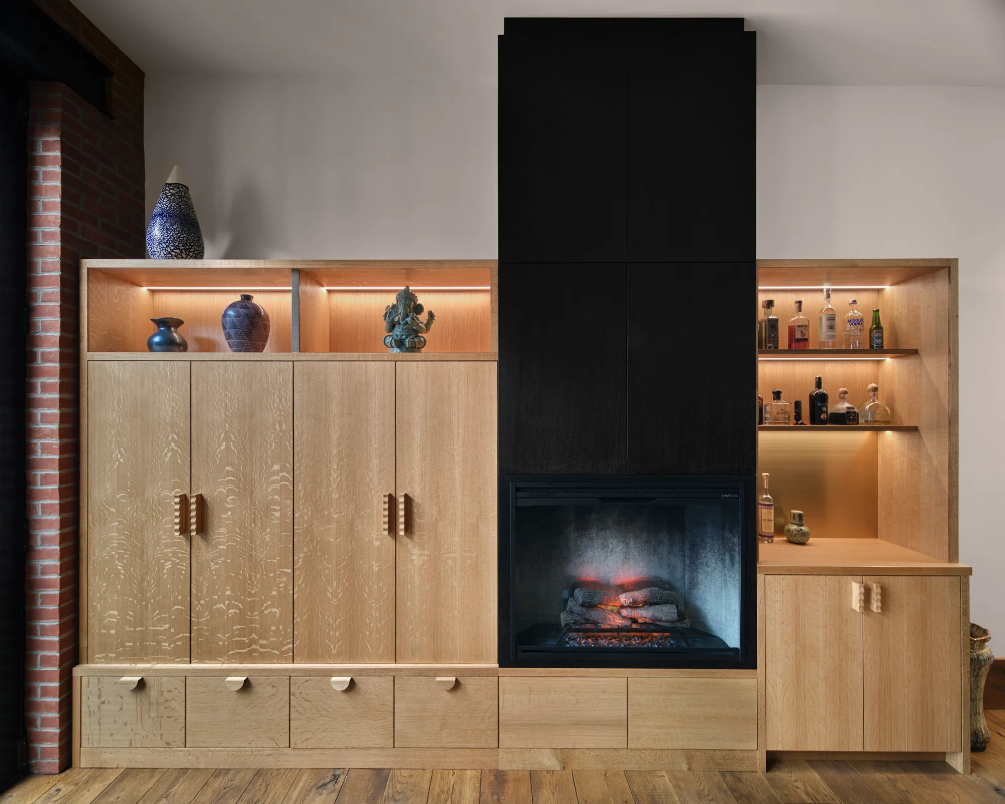 A modern wooden cabinet with open shelves and closed storage, decorated with vases, a sculpture, and alcohol bottles, and featuring a built-in electric fireplace.