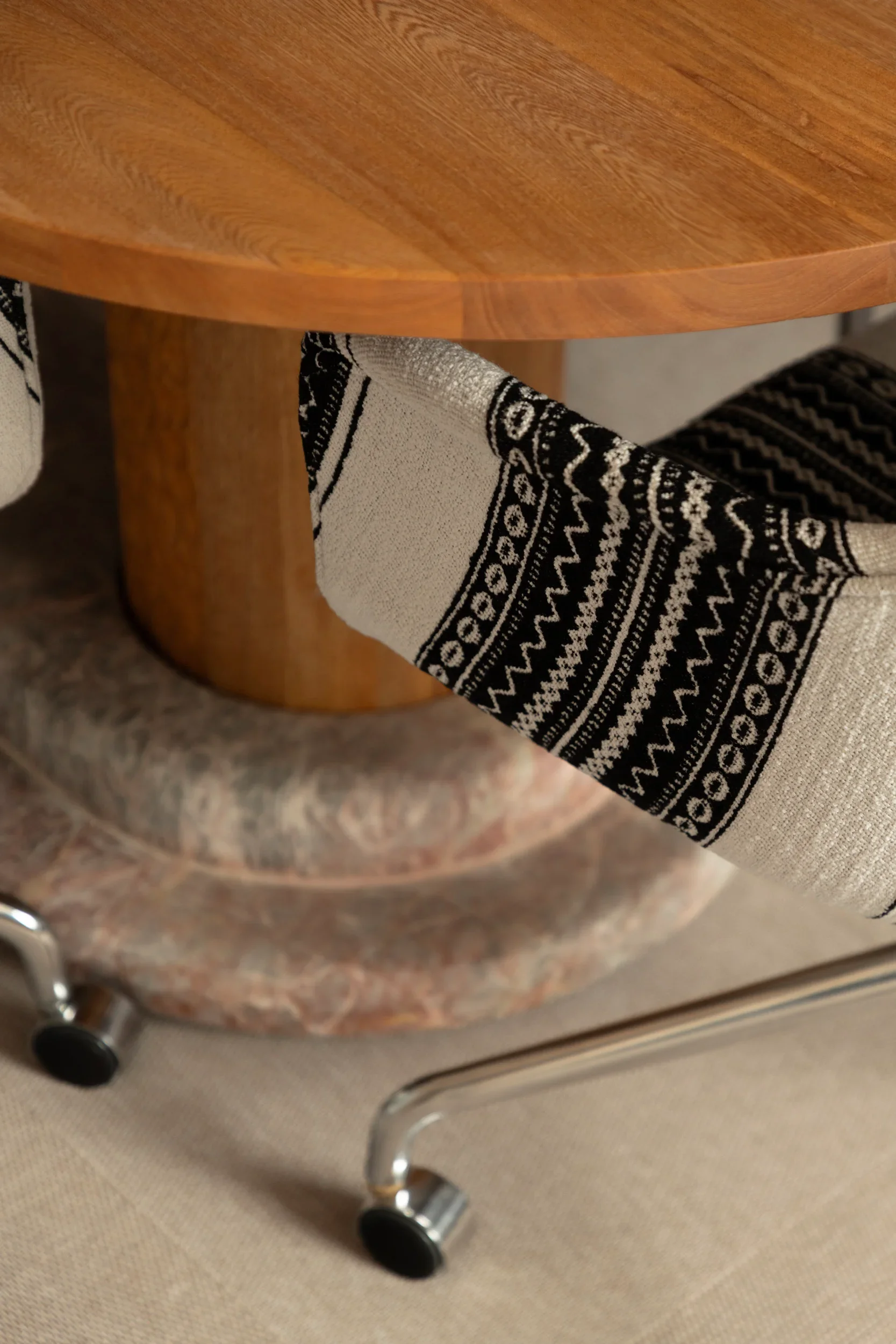 Close-up of a wooden round table with a fabric-covered chair underneath, set on a beige carpeted floor. The fabric has black and white patterns, and there's a metal chair leg with a black wheel visible in the bottom corner.